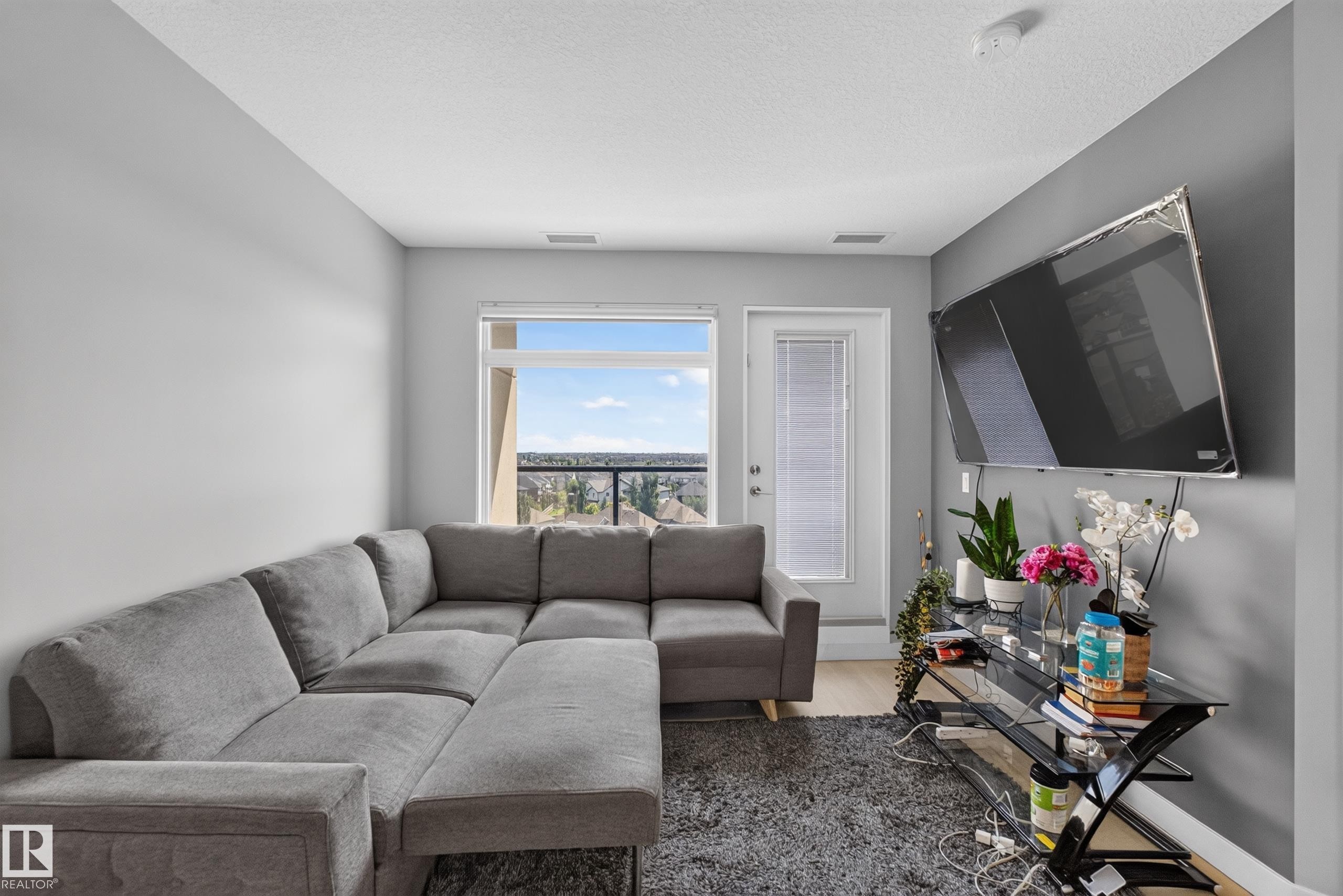 Living area featuring a large window, a glass-paneled balcony door, wood-finish flooring, and light gray walls - 706 5151 Windermere Boulevard, Edmonton, AB