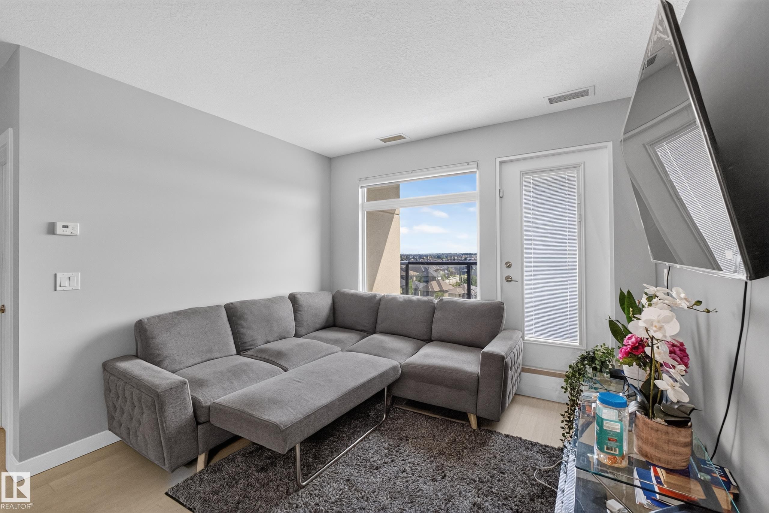 Living area featuring light wood-finish flooring, light grey walls, and a large window with an outdoor landscape - 706 5151 Windermere Boulevard, Edmonton, AB