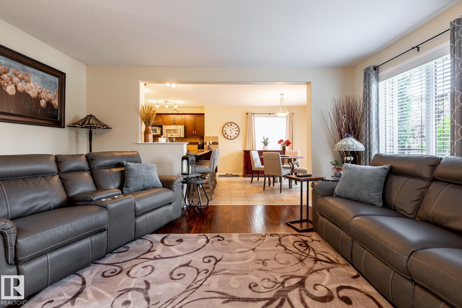 Living area with wood-finish flooring, a large window with blinds, and an open doorway to a dining area - 5820 7 Avenue Sw, Edmonton, AB - Indoor Photo Showing Living Room