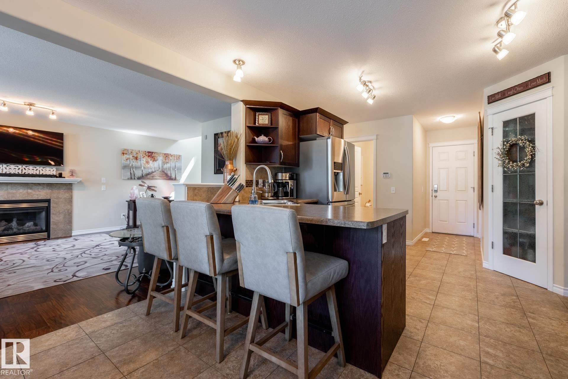 Open concept kitchen featuring dark wood cabinetry, a breakfast bar with seating, a stainless steel refrigerator, and tile flooring - 5820 7 Avenue Sw, Edmonton, AB - Indoor With Fireplace