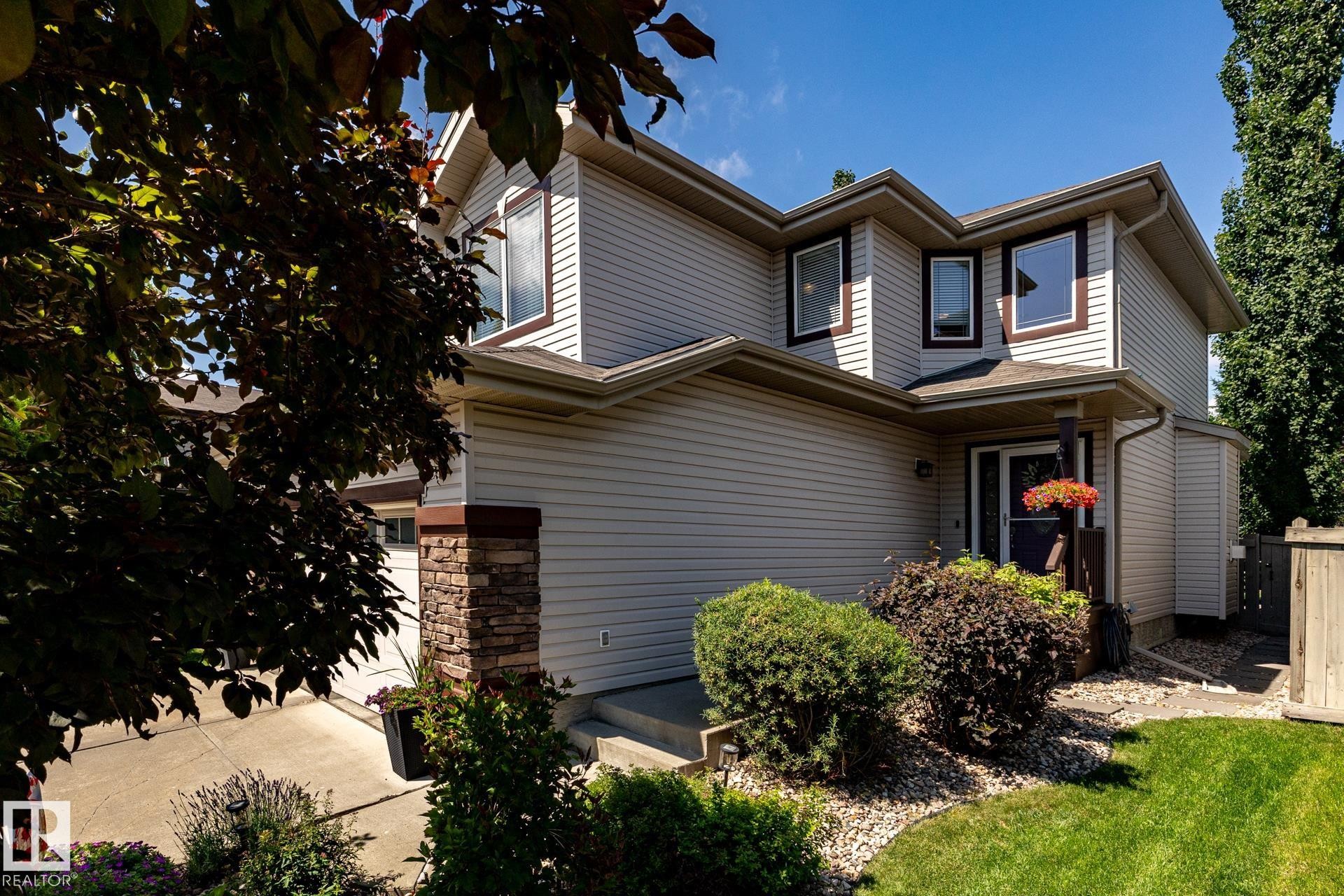 Two-story residence featuring light-toned siding, dark window trim, a stone-clad pillar, an integrated garage, and landscaped front yard with grass and shrubbery - 5820 7 Avenue Sw, Edmonton, AB - Outdoor
