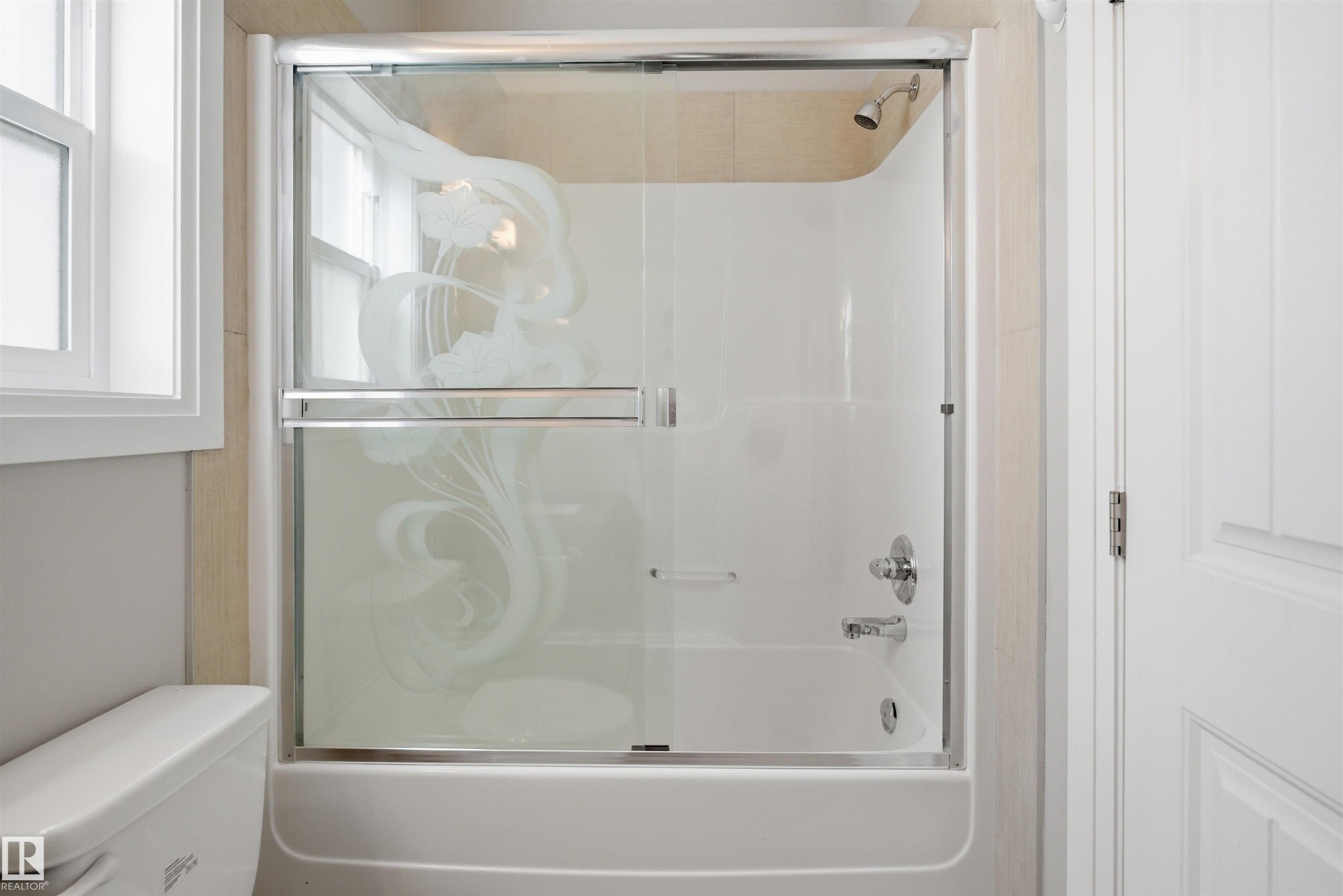 Bathtub and shower combination featuring a sliding glass enclosure with etched design, chrome fixtures, and light-toned tile surround - 16807 54 Street, Edmonton, AB - Indoor Photo Showing Bathroom