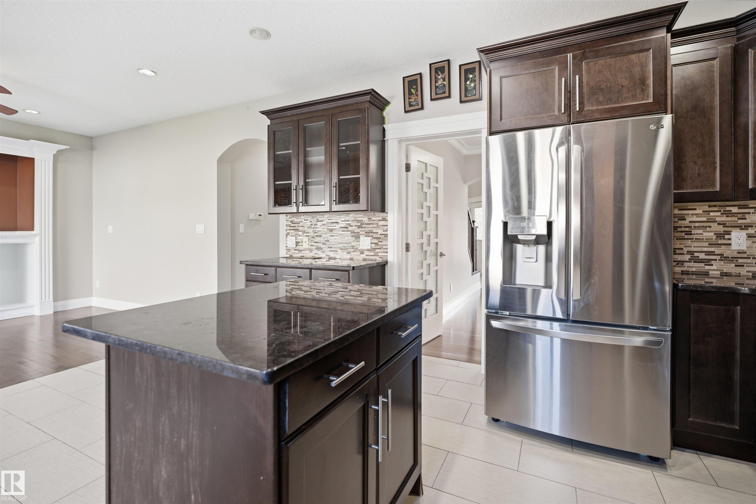 Kitchen featuring a central island with a dark stone countertop, dark wood cabinetry with glass-front uppers, a mosaic tile backsplash, and a stainless steel French door refrigerator - 16807 54 Street, Edmonton, AB - Indoor Photo Showing Kitchen With Stainless Steel Kitchen