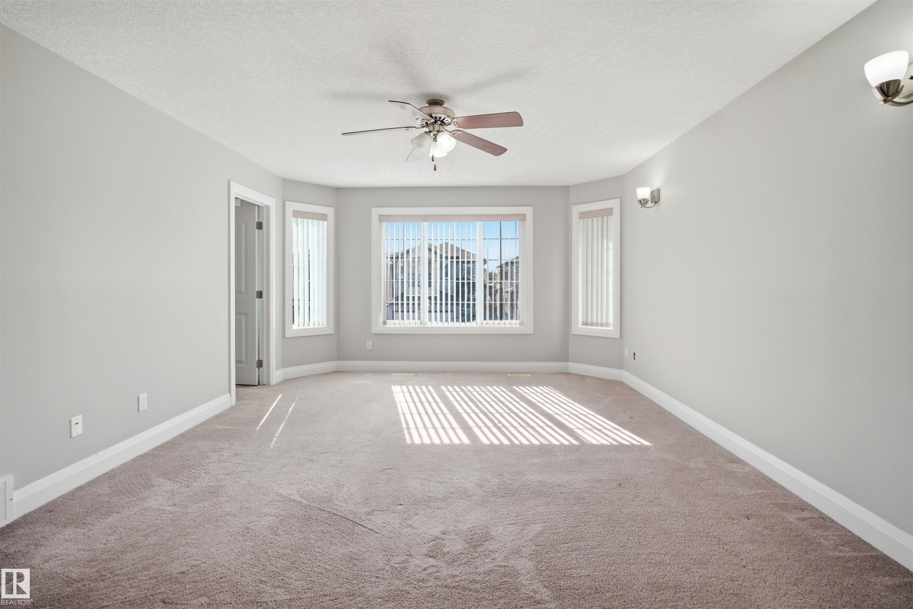 Expansive carpeted room featuring a bay window with vertical blinds, a ceiling fan with integrated lighting, and two wall-mounted sconces - 16807 54 Street, Edmonton, AB - Indoor Photo Showing Other Room