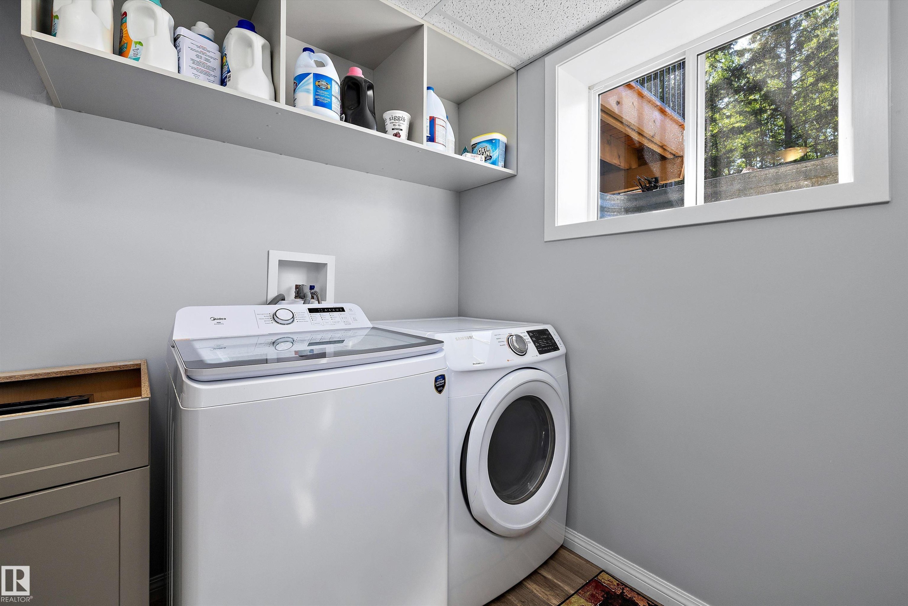 Dedicated laundry area featuring a white-framed window, overhead shelving, light gray wall paint, wood-finish flooring, and a built-in base cabinet - 53257 Rge Road 211, Rural Strathcona County, AB - Indoor Photo Showing Laundry Room