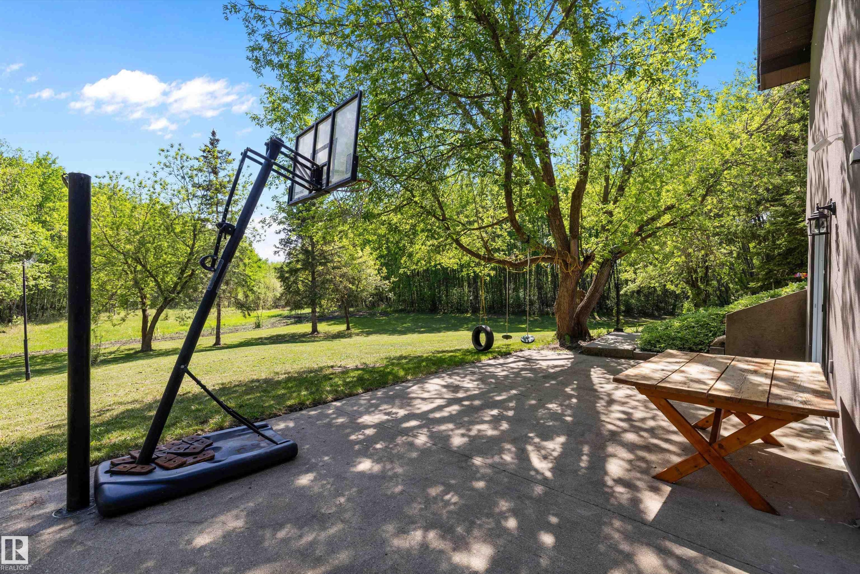 Expansive concrete patio offering a basketball hoop, mature trees, and a wooden outdoor table - 53257 Rge Road 211, Rural Strathcona County, AB - Outdoor