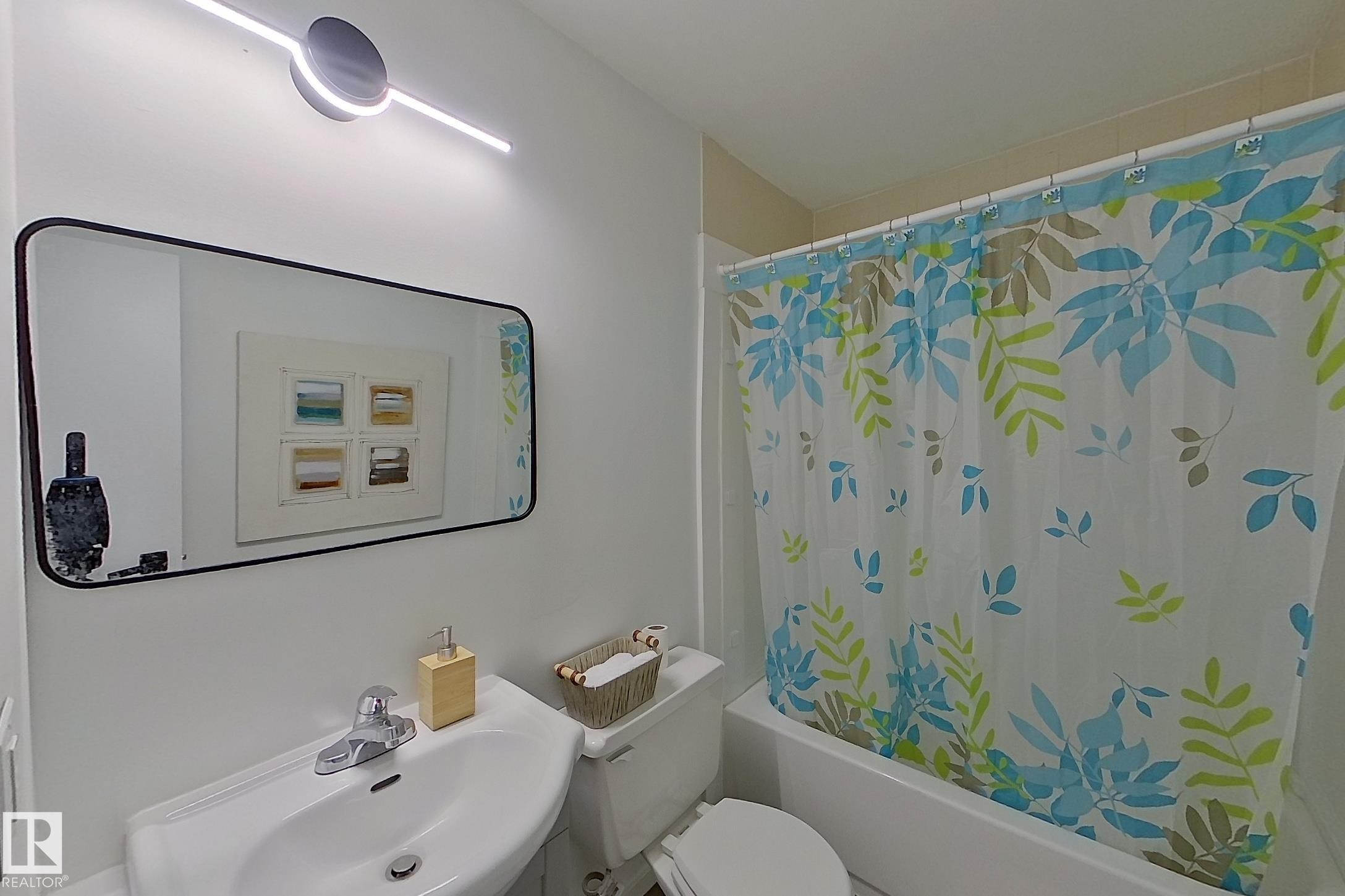 Bathroom featuring a white pedestal sink with chrome faucet, a rectangular mirror, and a white bathtub with a shower curtain - 209 Dickinsfield Court Nw, Edmonton, AB