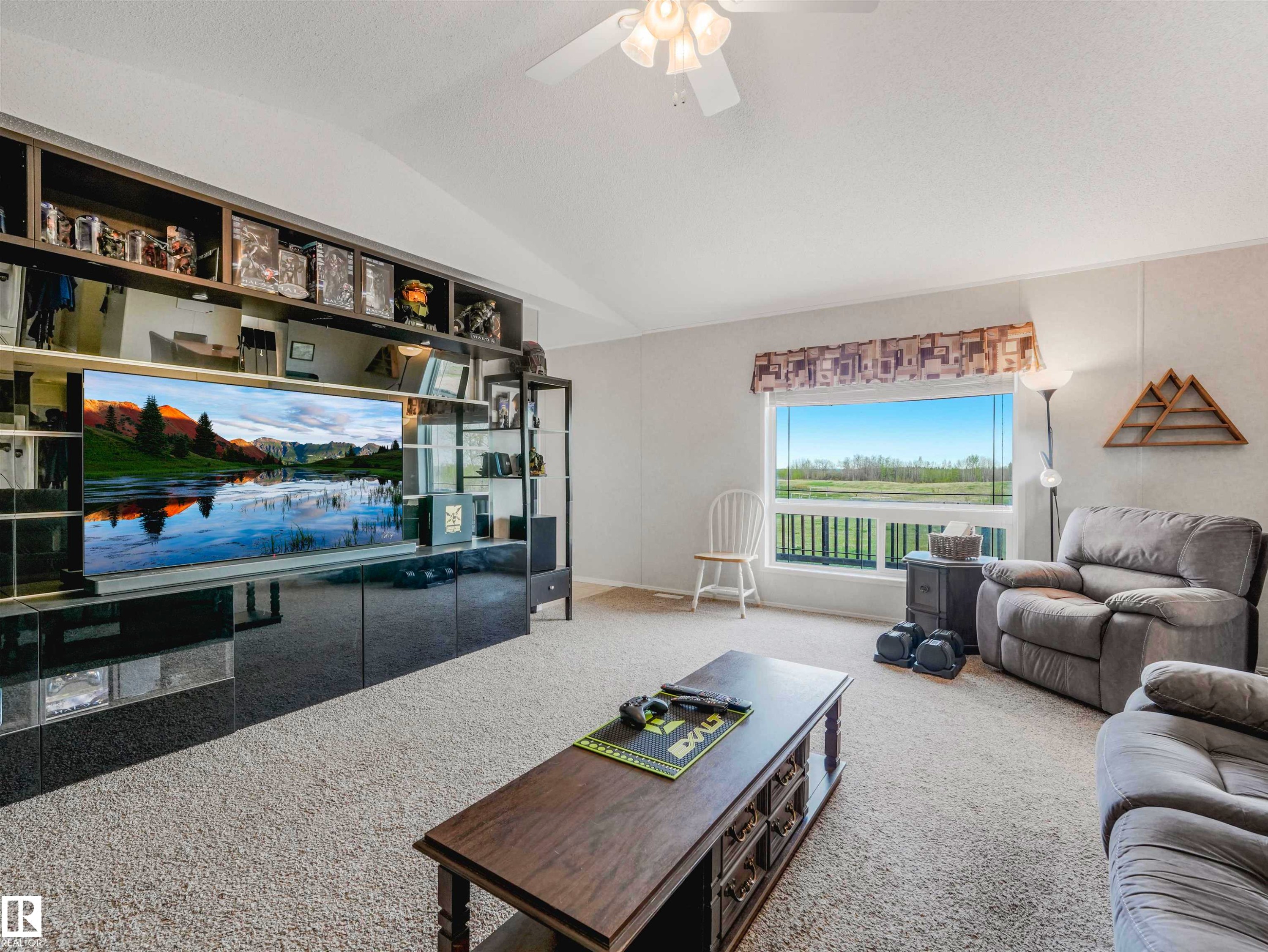 Carpeted living area featuring a vaulted ceiling with a ceiling fan, a large window, and a built-in entertainment center with open shelving and lower cabinetry - 57016 Range Road 13, Rural Barrhead County, AB - Indoor Photo Showing Living Room