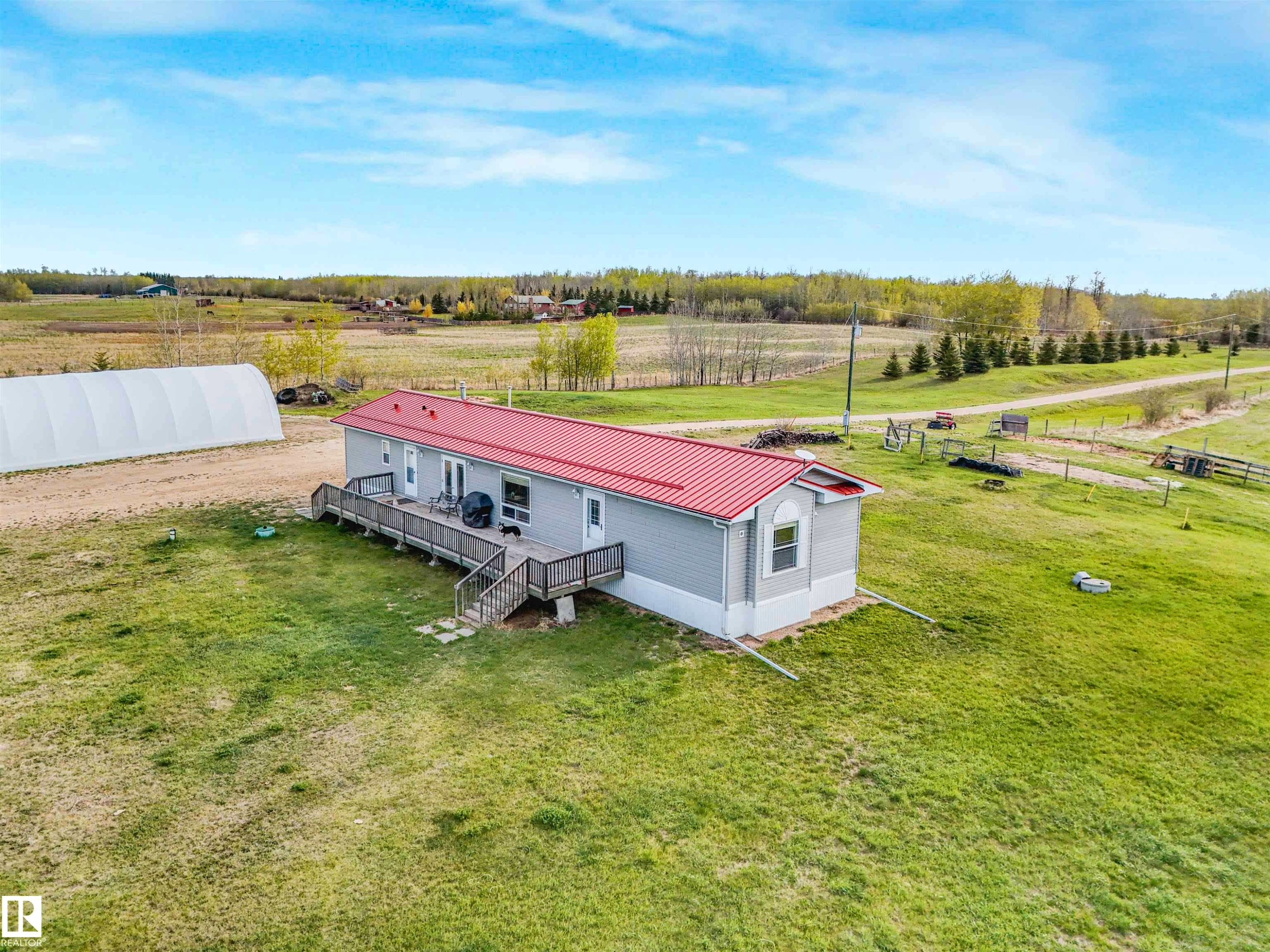 Single-story residence featuring a red metal roof, light gray siding, and a spacious wood-finish deck with railings - 57016 Range Road 13, Rural Barrhead County, AB - Outdoor With View