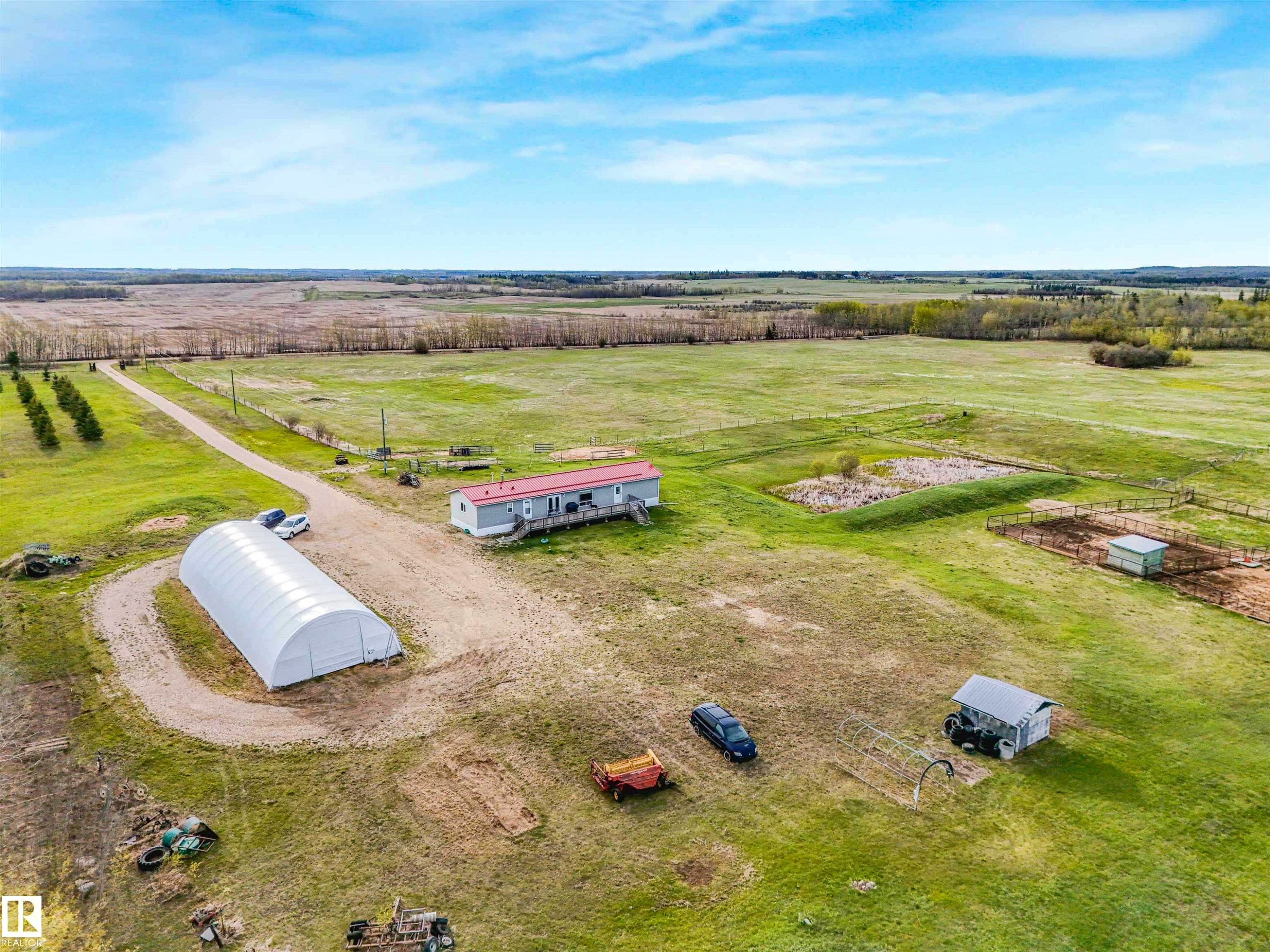 Expansive rural property featuring a long gravel driveway, a single-story residence with a covered porch, a large white hoop structure, a fenced animal enclosure, and multiple outbuildings across open fields - 57016 Range Road 13, Rural Barrhead County, AB - Outdoor With View