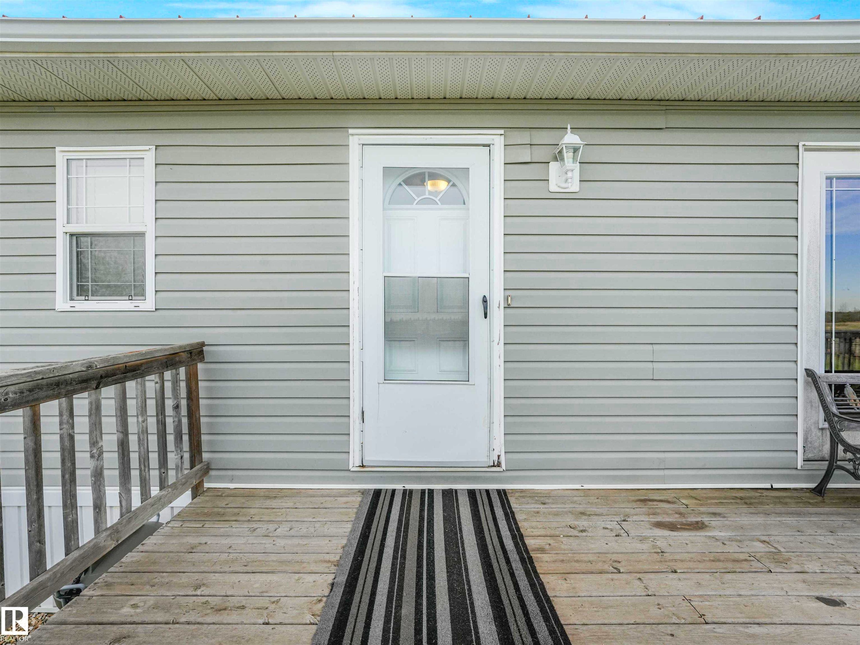 Light grey horizontal siding exterior featuring a white paneled entry door with a storm door, a white exterior light fixture, and a wooden ramp leading to the entrance - 57016 Range Road 13, Rural Barrhead County, AB - Outdoor With Exterior