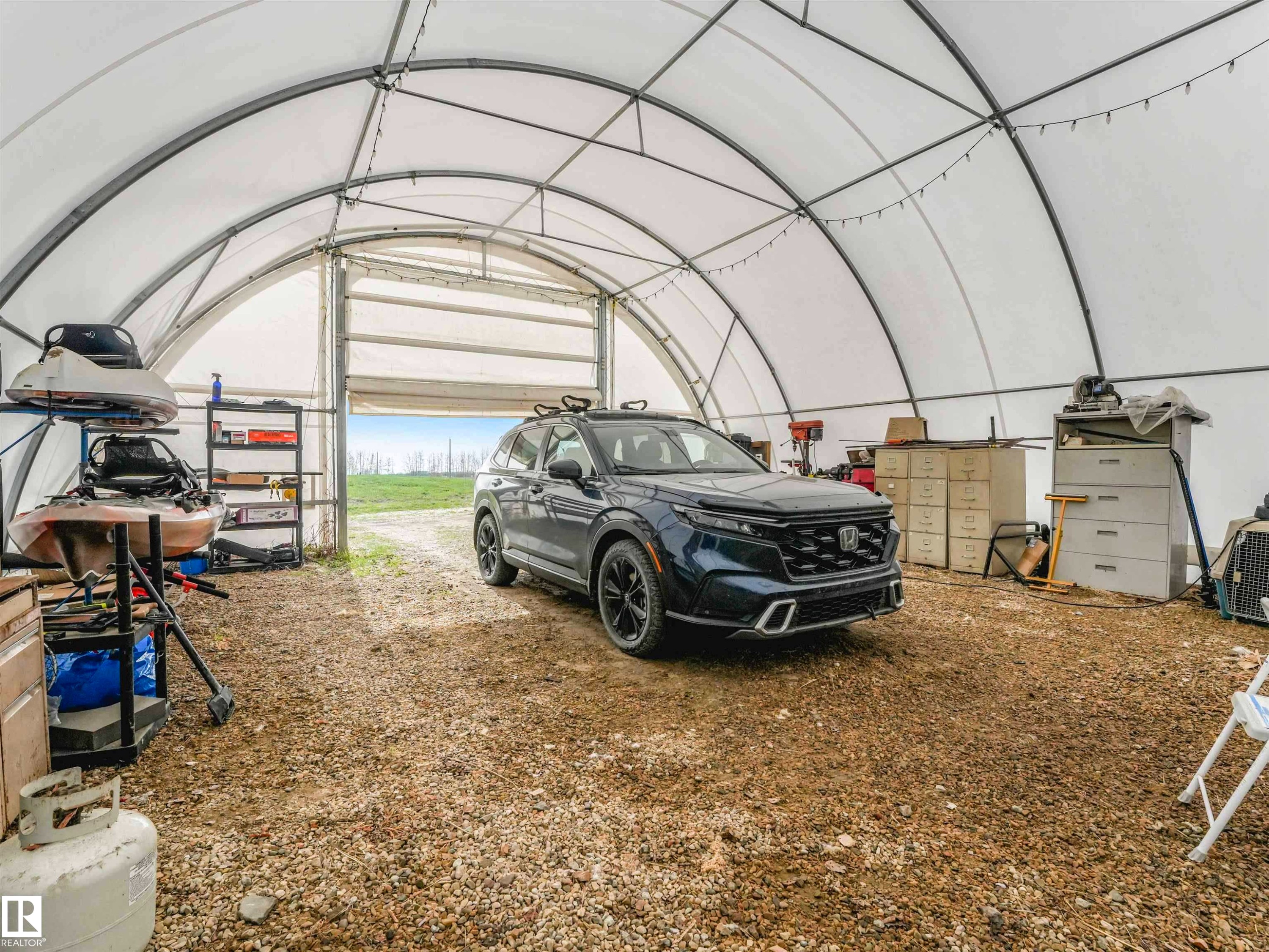 Spacious arched structure featuring a roll-up door, gravel flooring, interior lighting, and exposed metal framing - 57016 Range Road 13, Rural Barrhead County, AB - Indoor Photo Showing Garage