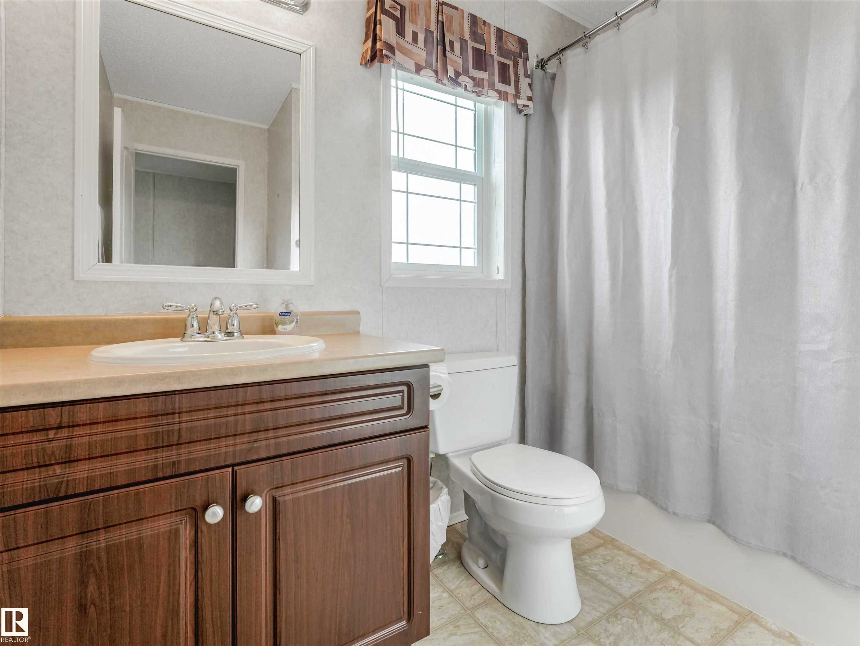 Bathroom featuring a wood-finish vanity with a light-toned countertop, a rectangular mirror, a white toilet, and a window with decorative valance - 57016 Range Road 13, Rural Barrhead County, AB - Indoor Photo Showing Bathroom