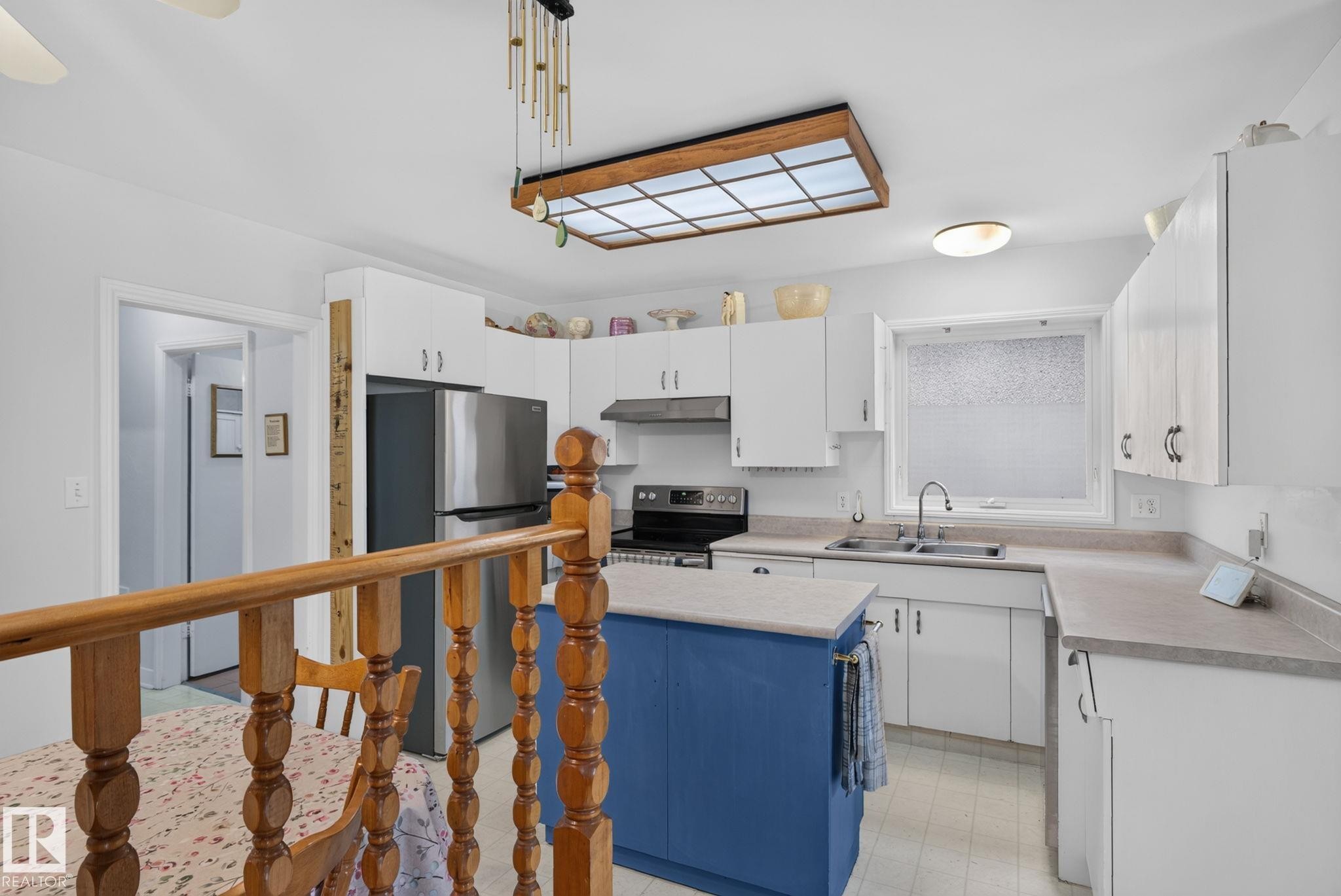 Bright kitchen featuring white cabinetry, a central blue island, stainless steel refrigerator, and a double basin sink - 12703 122 Street Nw, Edmonton, AB