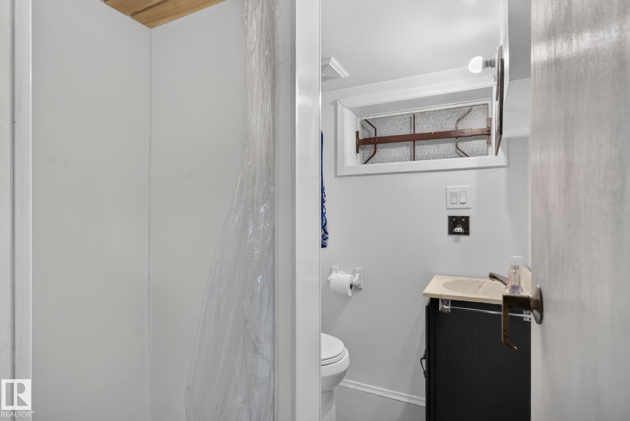 Bathroom featuring a white toilet, black vanity with a light-colored countertop, and a shower area with white walls - 12703 122 Street Nw, Edmonton, AB