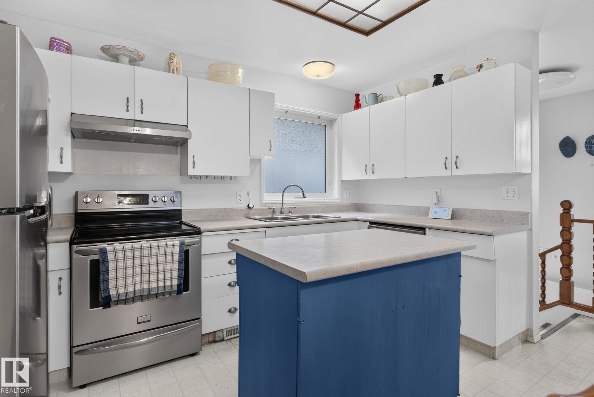 Bright kitchen featuring white cabinetry, light-toned countertops, stainless steel appliances, and a central island with a blue-painted base - 12703 122 Street Nw, Edmonton, AB