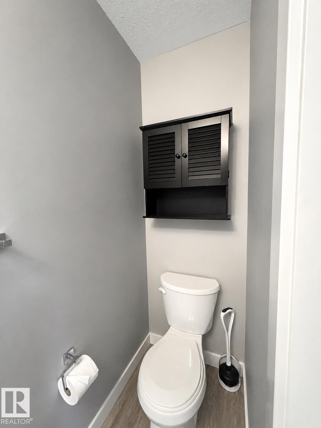 Compact bathroom featuring a white toilet, wood-finish flooring, white baseboards, and a black wall-mounted cabinet with louvered doors - 187 Radcliffe Wynd, Fort Saskatchewan, AB - Indoor Photo Showing Bathroom