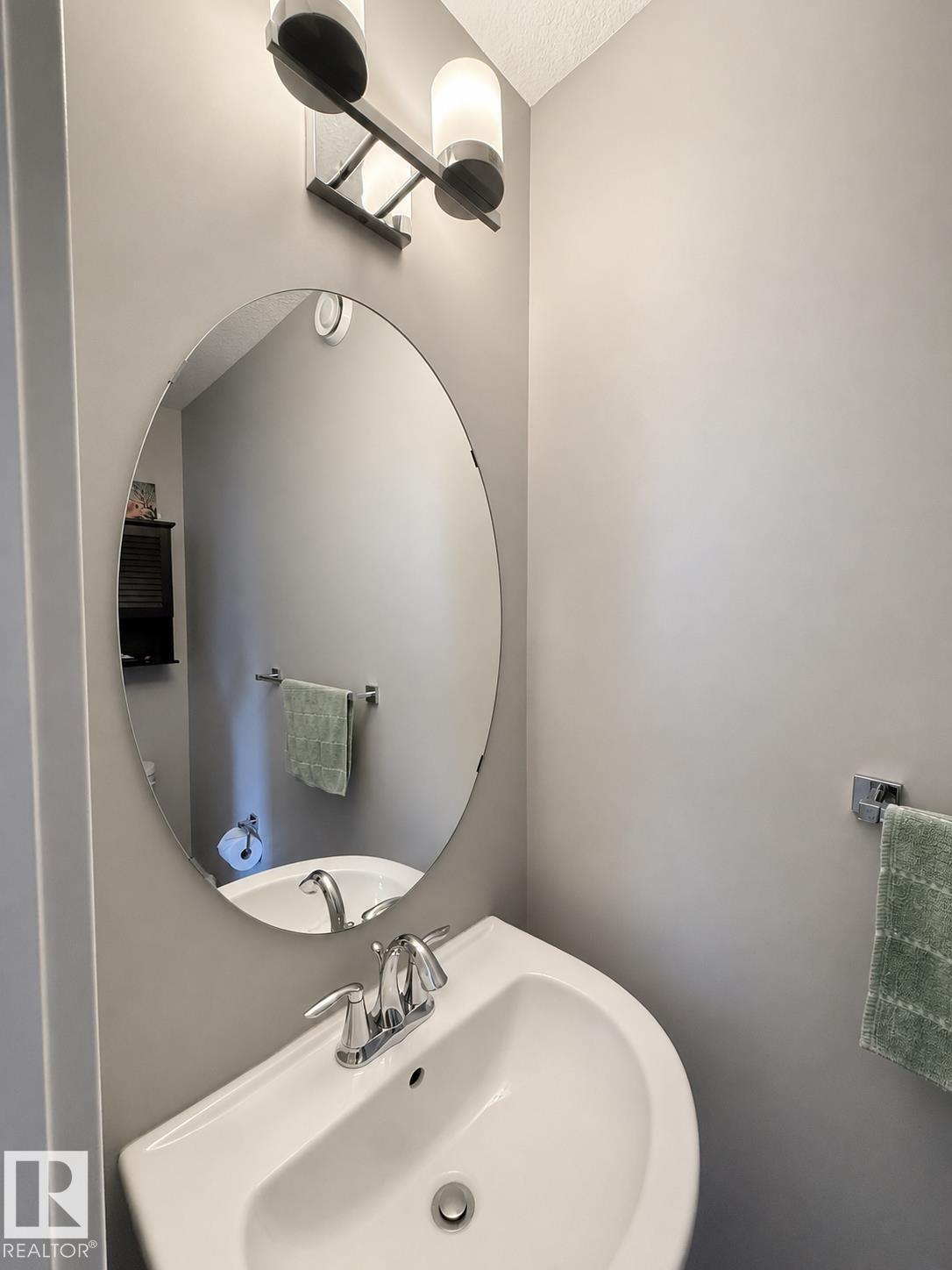 Pedestal sink with chrome faucet, complemented by an oval wall mirror and modern vanity lighting - 187 Radcliffe Wynd, Fort Saskatchewan, AB - Indoor Photo Showing Bathroom