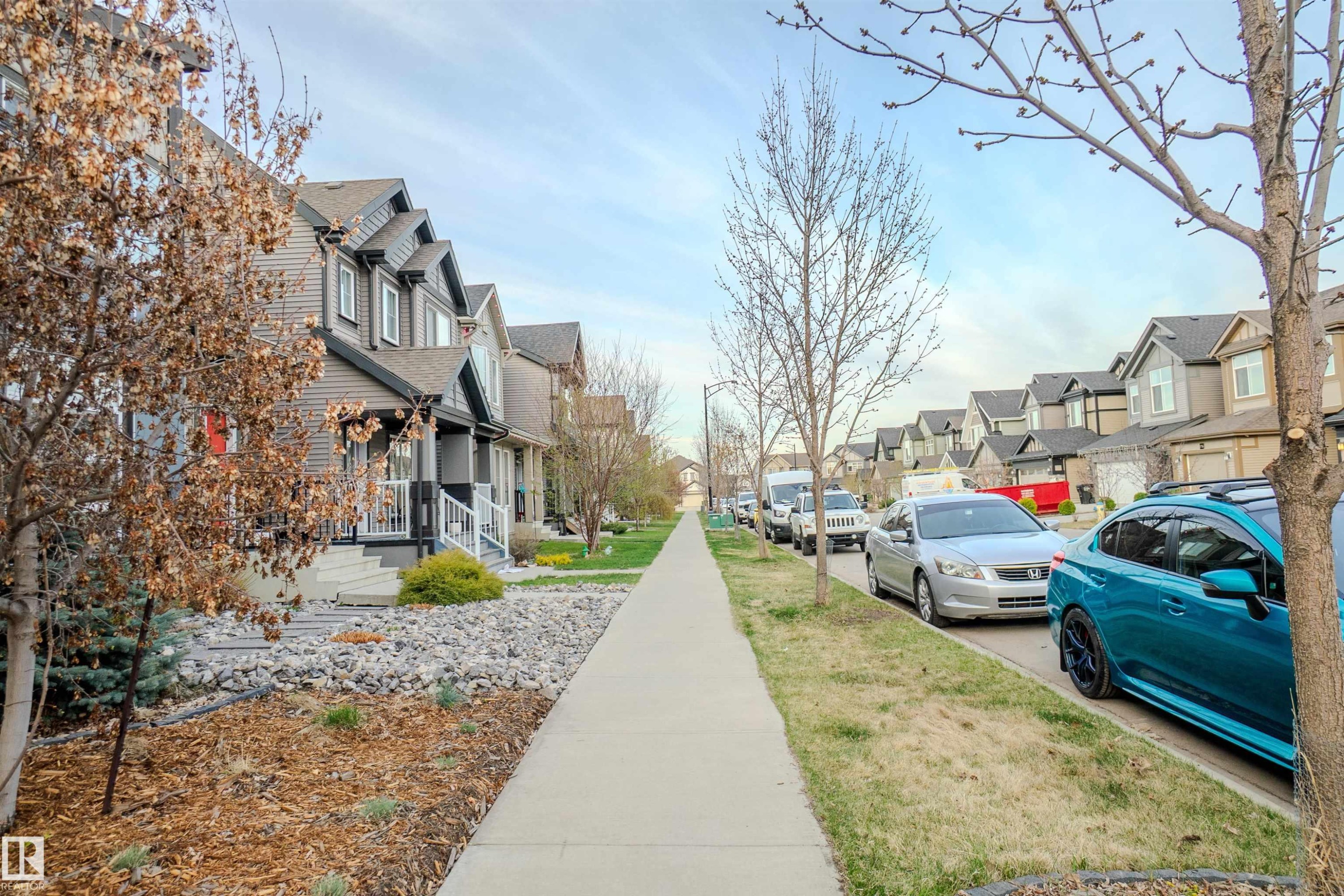 Street scene featuring a concrete sidewalk, mature trees, and a row of residential properties with varied siding, front porches, and gabled roofs - 1766 Dumont Crescent Sw, Edmonton, AB - Outdoor