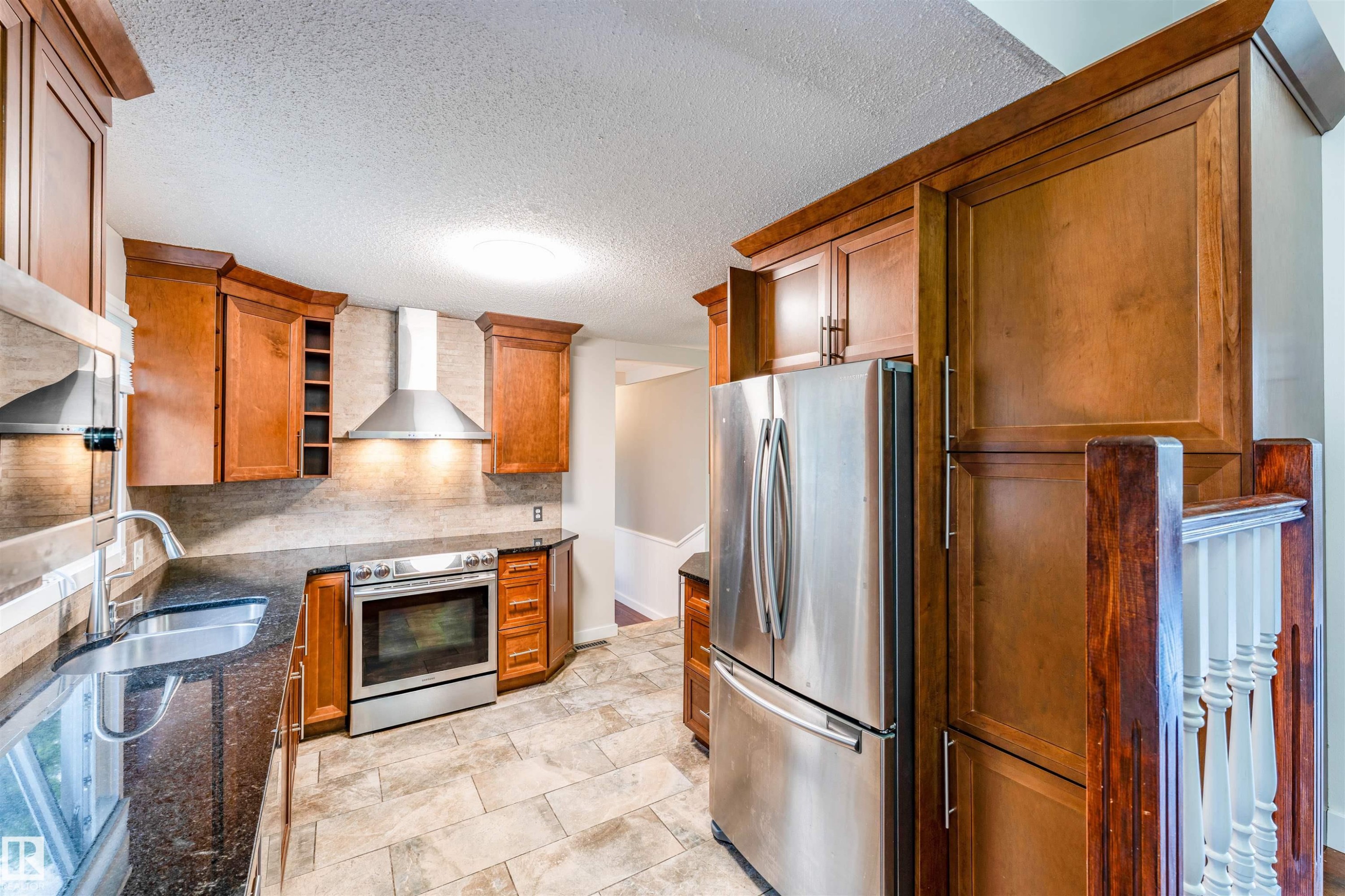 Kitchen featuring rich wood cabinetry, stainless steel appliances, dark stone countertops, tile backsplash, and tile flooring - 5212 142 Street Nw, Edmonton, AB