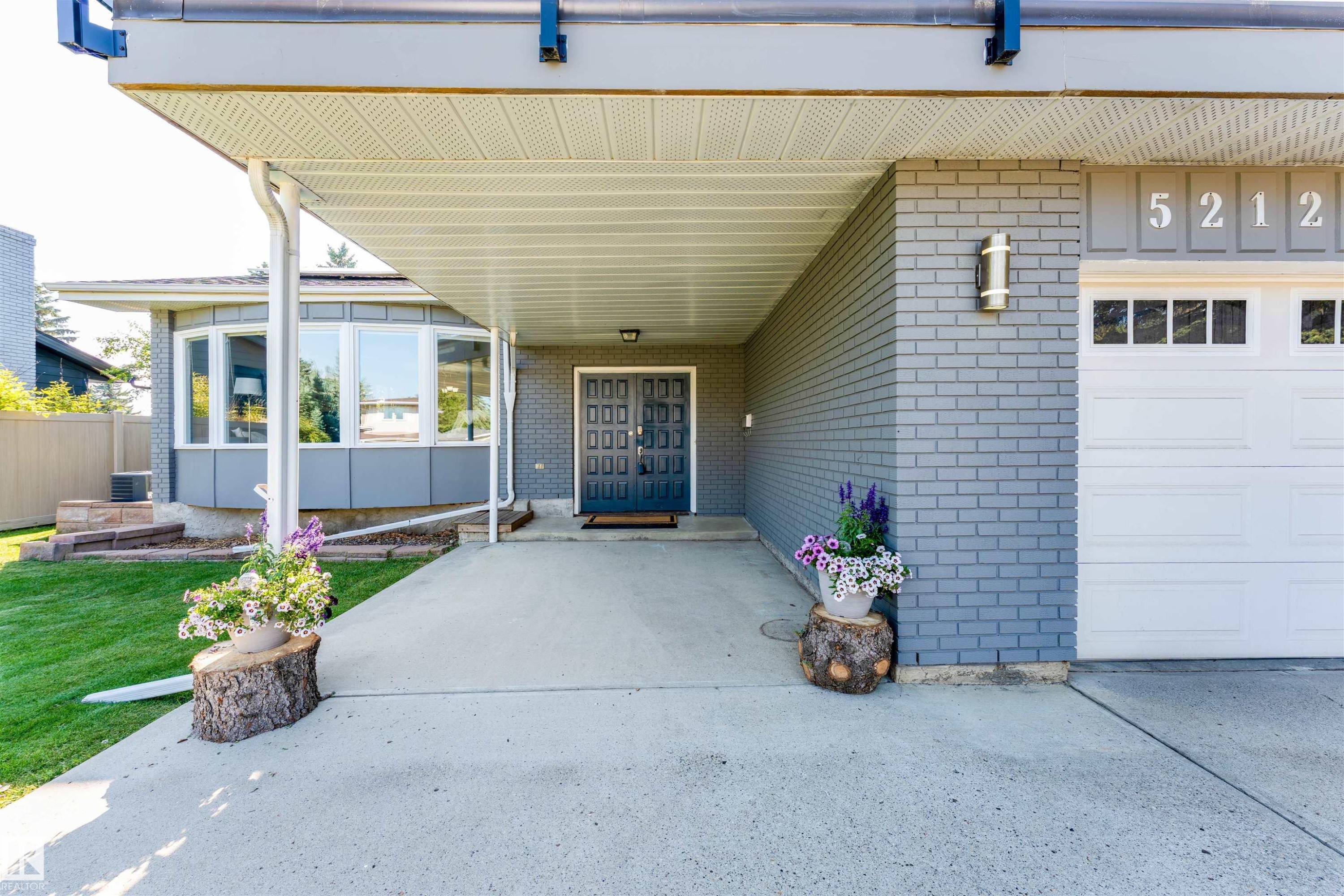 Covered entryway featuring a bay window, concrete driveway, attached garage with window inserts, and grey brick exterior - 5212 142 Street Nw, Edmonton, AB