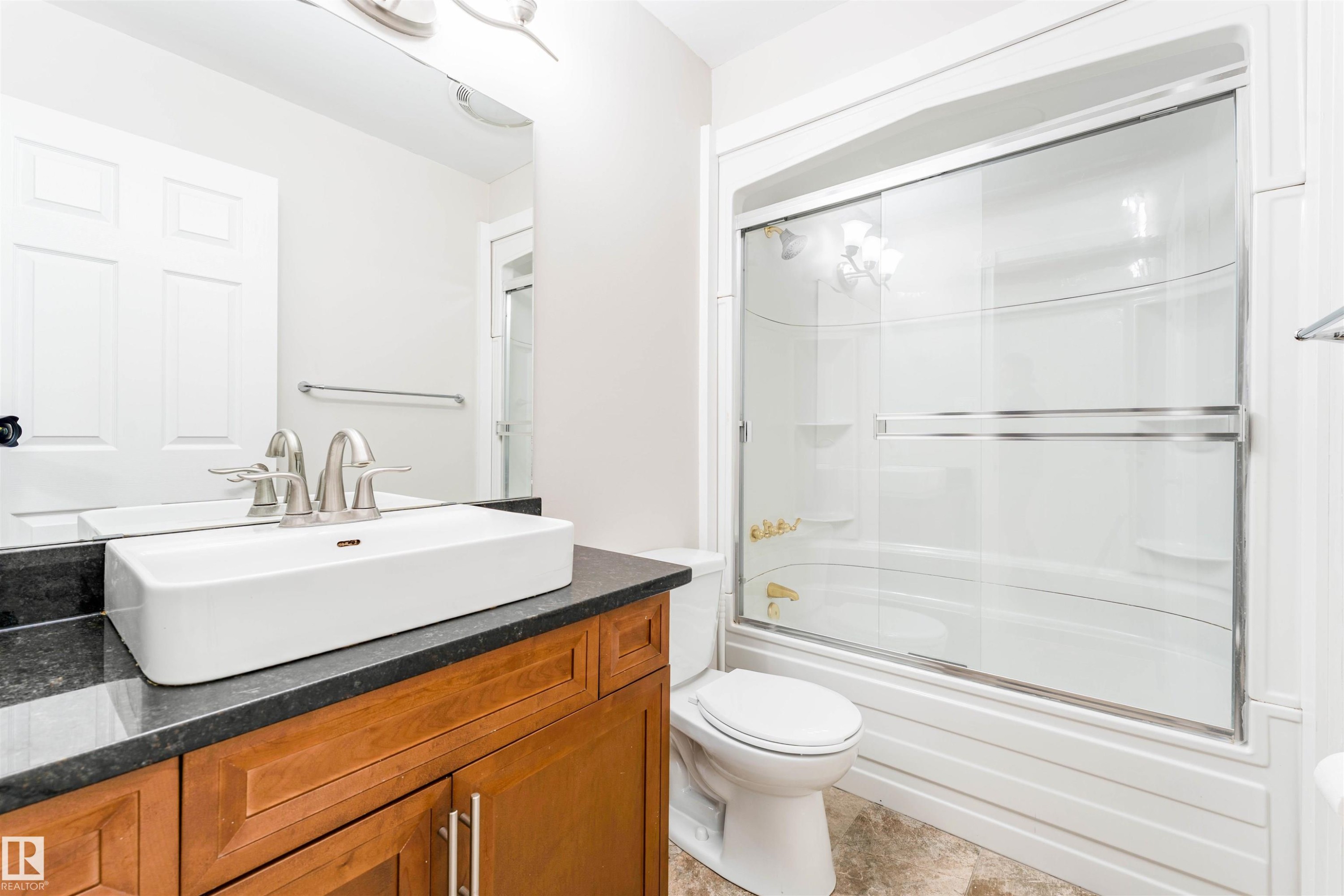 Bathroom featuring a rectangular vessel sink, dark countertop, wood-finish vanity, and a shower-tub combination with sliding glass doors - 5212 142 Street Nw, Edmonton, AB