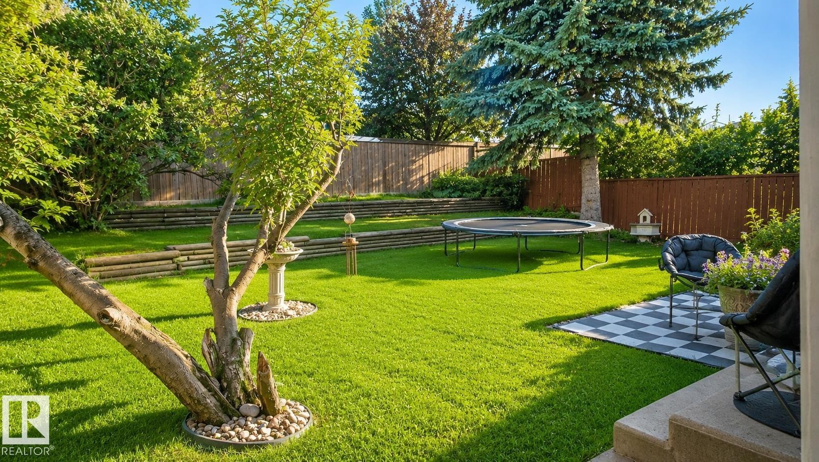 Spacious backyard featuring a lush lawn, mature trees, tiered landscaping with wood retaining walls, and a checkered patio area - 1022 106 Street Nw, Edmonton, AB - Outdoor With Backyard