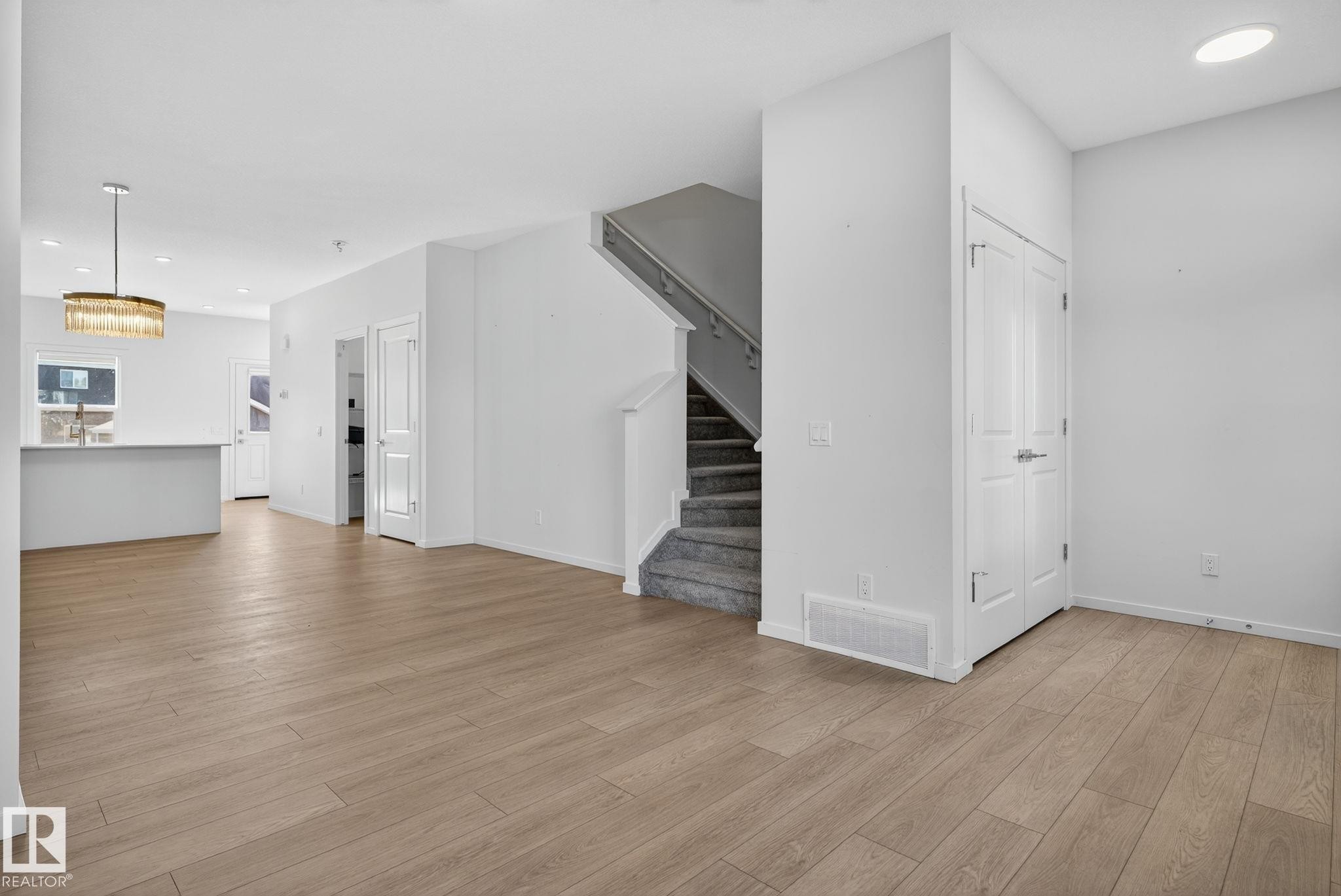 Open-concept living area featuring wood-finish flooring, a contemporary chandelier, a carpeted staircase, and white bi-fold doors - 264 Orchards Boulevard Sw, Edmonton, AB - Indoor
