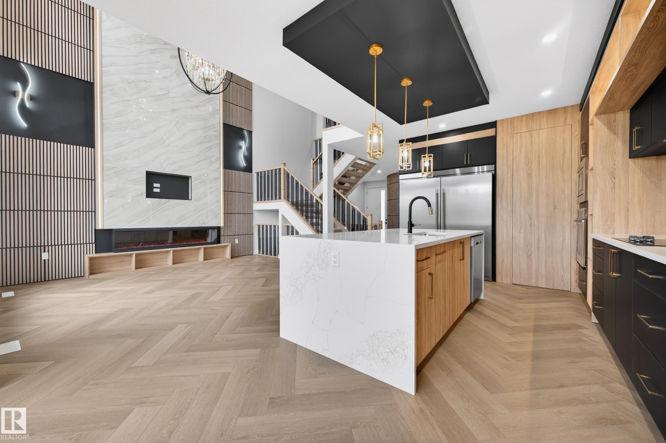 Open-concept living space featuring herringbone wood-finish flooring, a contemporary kitchen island with a white countertop and wood-finish cabinetry, and a striking fireplace wall with marble-finish paneling and wood slat accents - 431 172 Avenue, Edmonton, AB - Indoor Photo Showing Kitchen