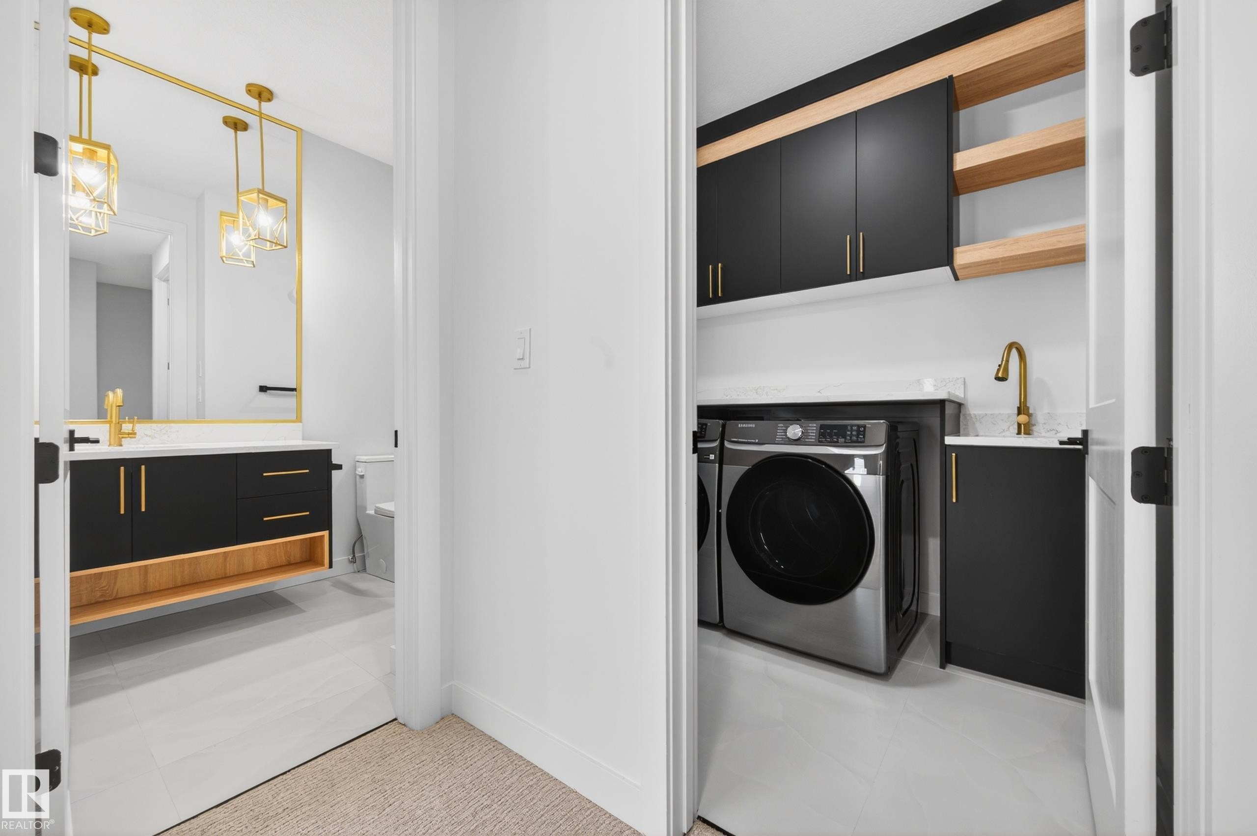 Modern laundry area featuring a built-in sink, front-load washer and dryer, dark cabinetry with gold-tone hardware, and open wood shelving - 431 172 Avenue, Edmonton, AB - Indoor Photo Showing Laundry Room