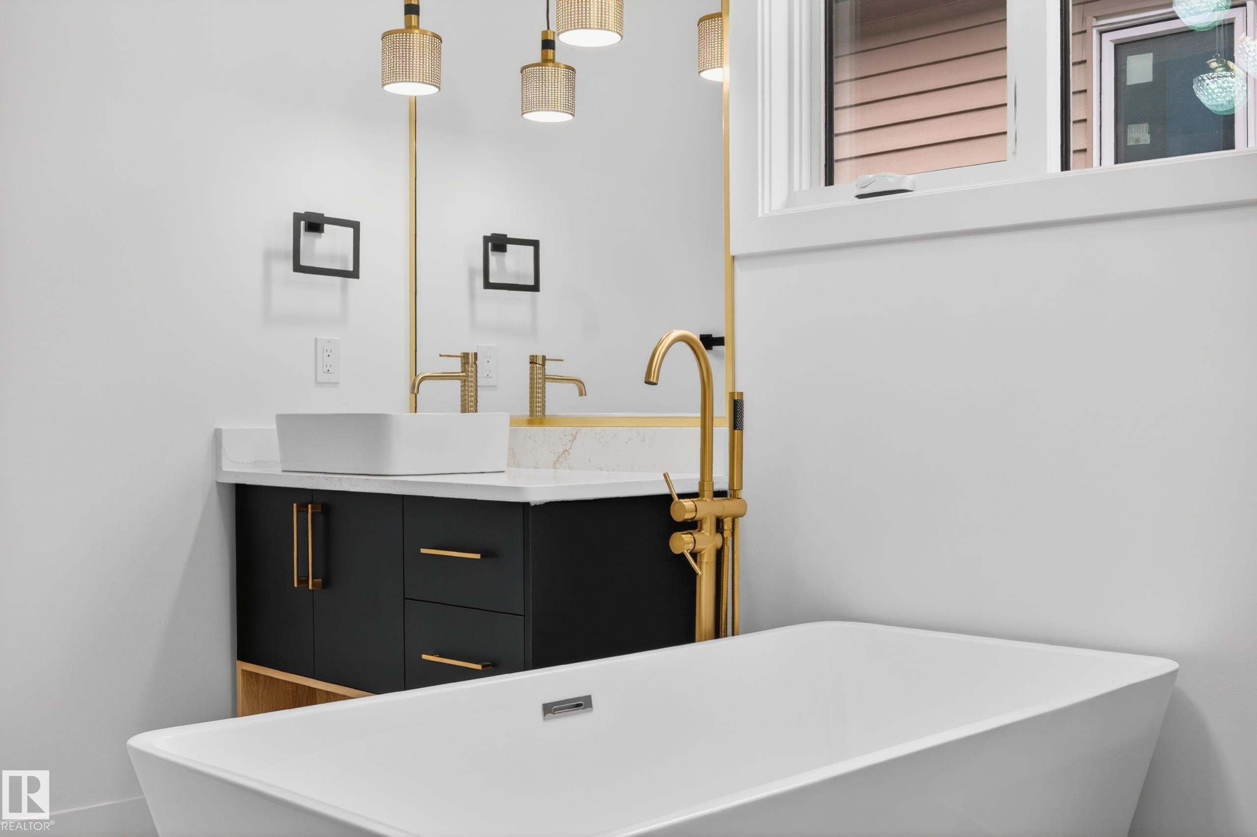Freestanding soaking tub with a floor-mounted gold-tone faucet, accompanied by a dual vanity featuring black cabinetry, gold-tone hardware, and white rectangular vessel sinks - 431 172 Avenue, Edmonton, AB - Indoor