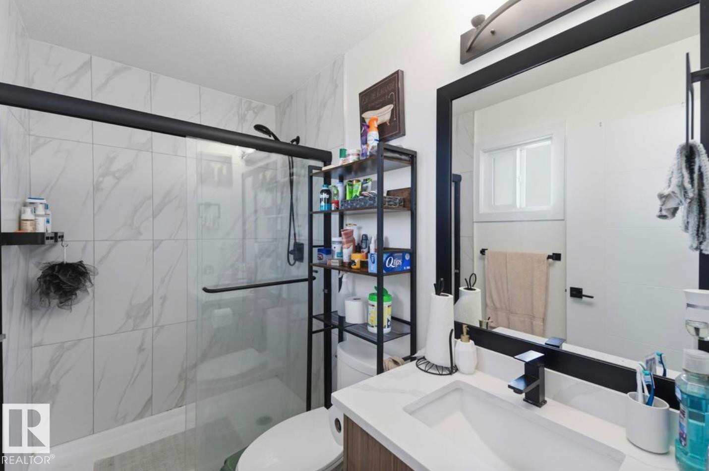 Bathroom featuring a glass-enclosed shower with white marble-look tile, a single vanity with a white rectangular sink, a black framed mirror, and matte black fixtures - 17924 61 Street Nw, Edmonton, AB - Indoor Photo Showing Bathroom