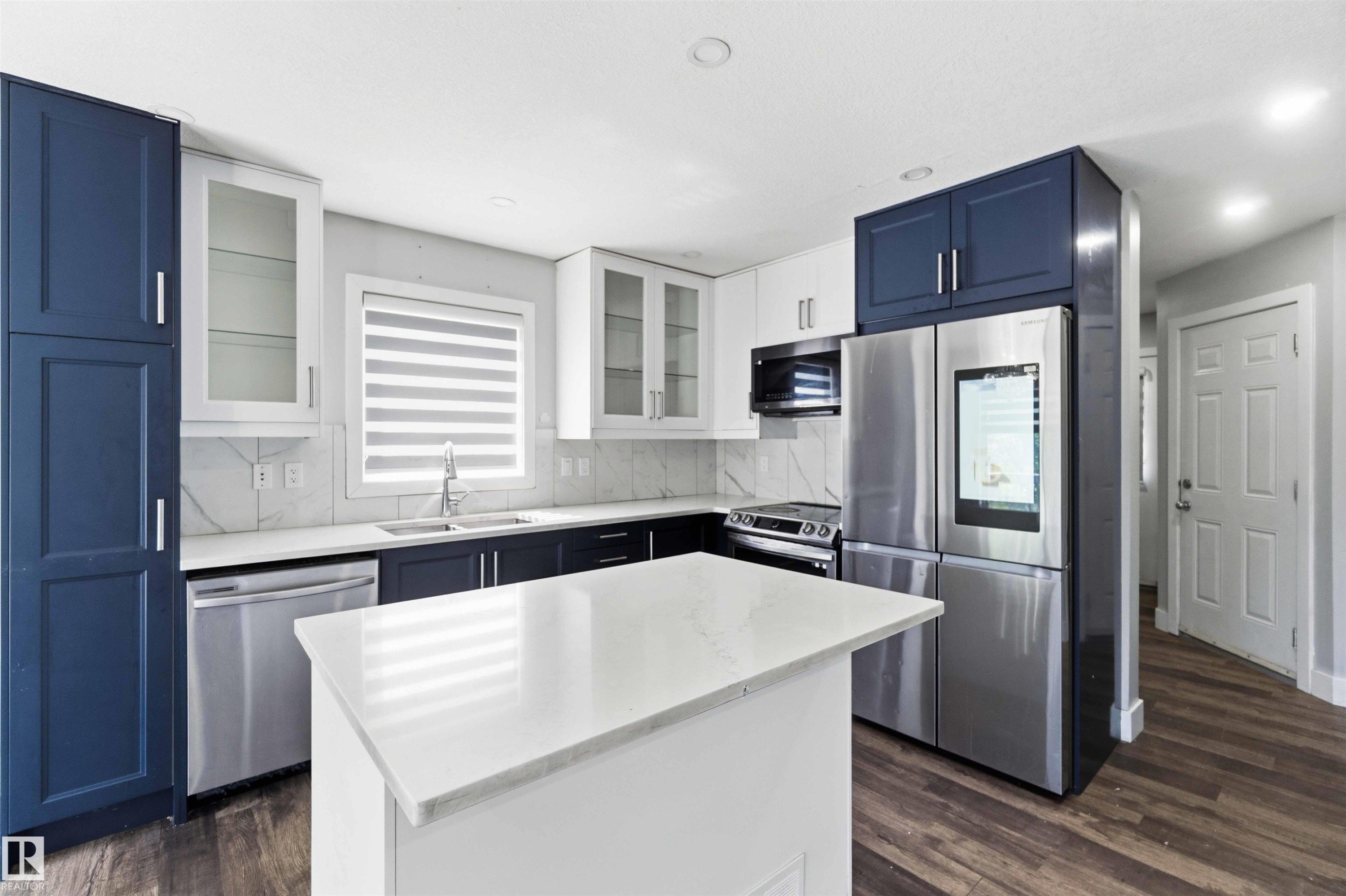 Kitchen featuring two-tone cabinetry with glass-front uppers, stainless steel appliances, a center island with a solid surface countertop, and wood-finish flooring - #64 120 Magrath Rd Nw, Edmonton, AB - Indoor Photo Showing Kitchen With Upgraded Kitchen