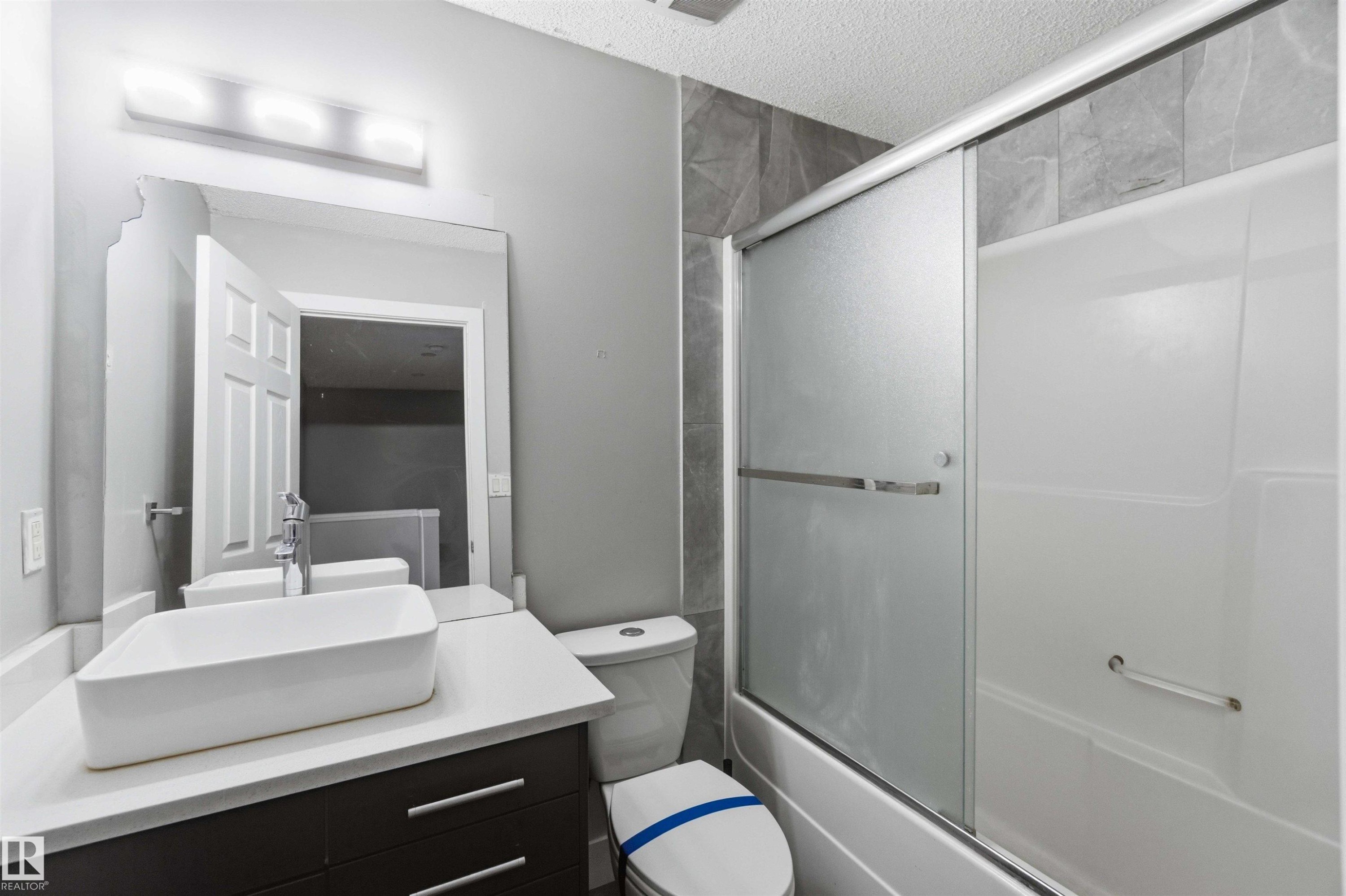 Modern bathroom featuring a white vessel sink on a light-colored countertop, a dark wood-finish vanity with drawer pulls, and a bathtub with sliding frosted glass doors - #64 120 Magrath Rd Nw, Edmonton, AB - Indoor Photo Showing Bathroom