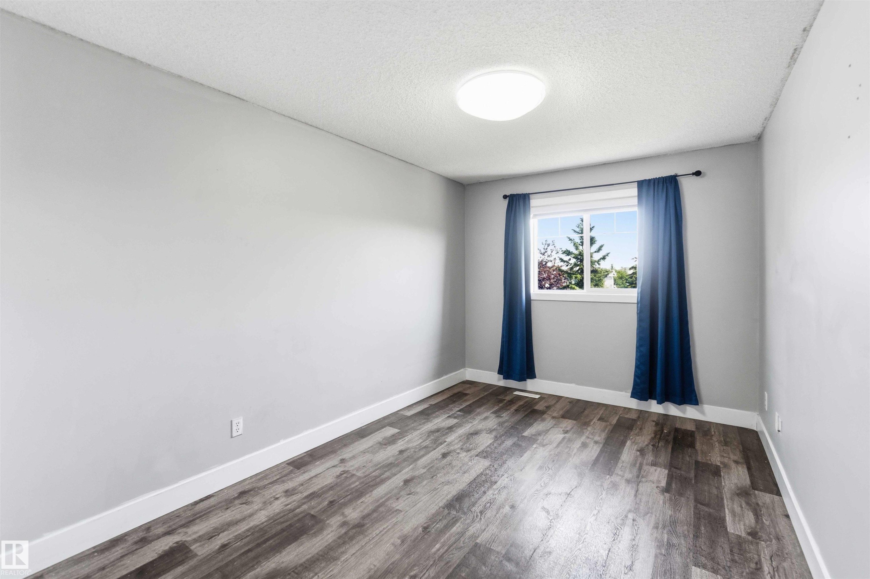 Room featuring wood-finish flooring, light gray walls, white baseboards, and a window with blue drapes and a black rod - #64 120 Magrath Rd Nw, Edmonton, AB - Indoor Photo Showing Other Room