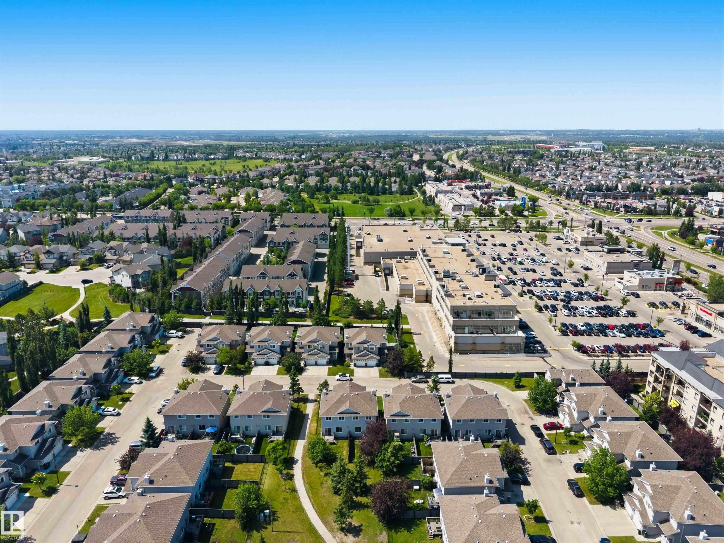 Expansive aerial view showcasing residential properties, a commercial complex with extensive parking, and a multi-lane roadway - #64 120 Magrath Rd Nw, Edmonton, AB - Outdoor With View
