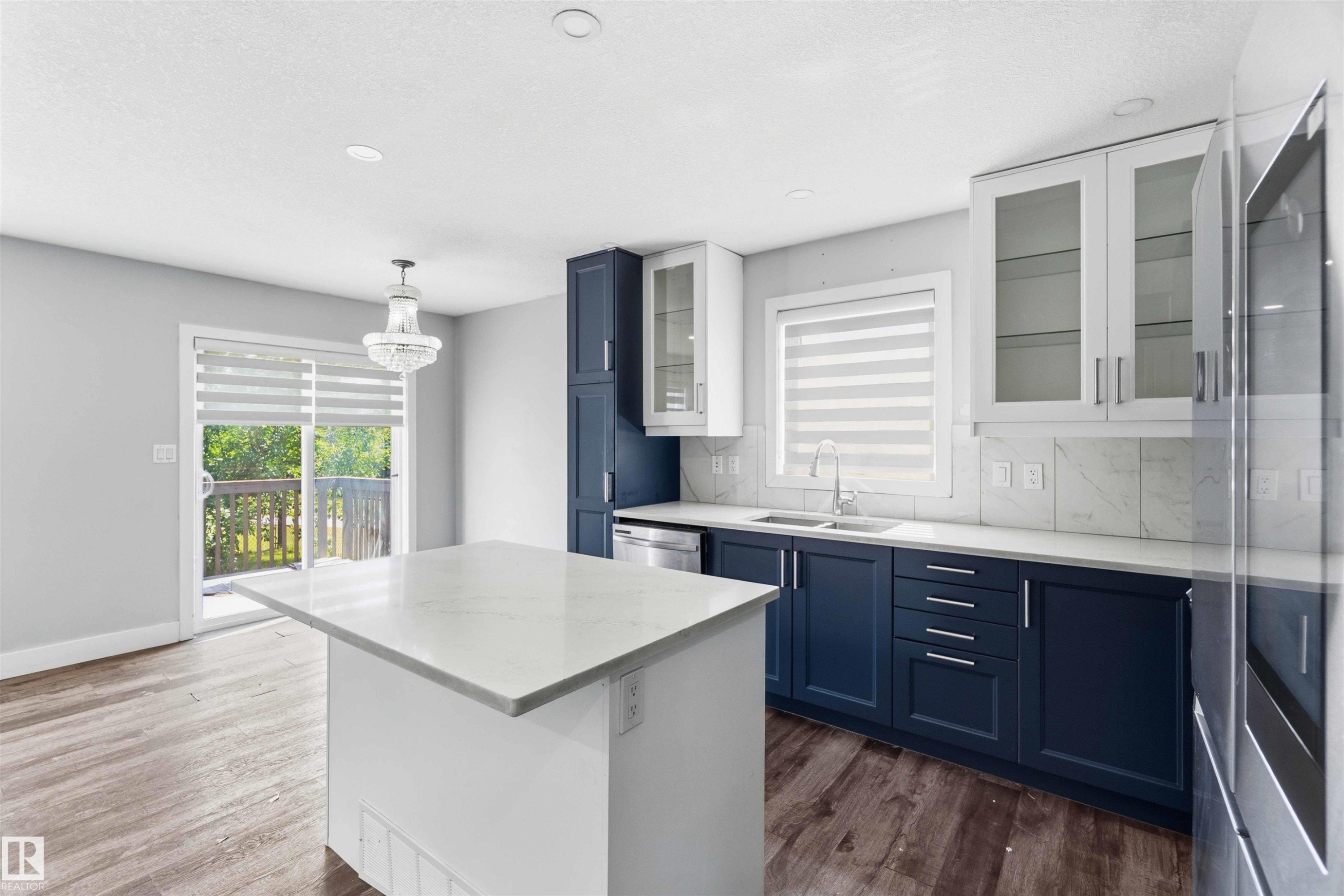 Contemporary kitchen featuring a central island with a light-toned countertop, two-tone cabinetry with dark lower and white upper units, a stainless steel sink with a modern faucet, and wood-finish flooring - #64 120 Magrath Rd Nw, Edmonton, AB - Indoor Photo Showing Kitchen