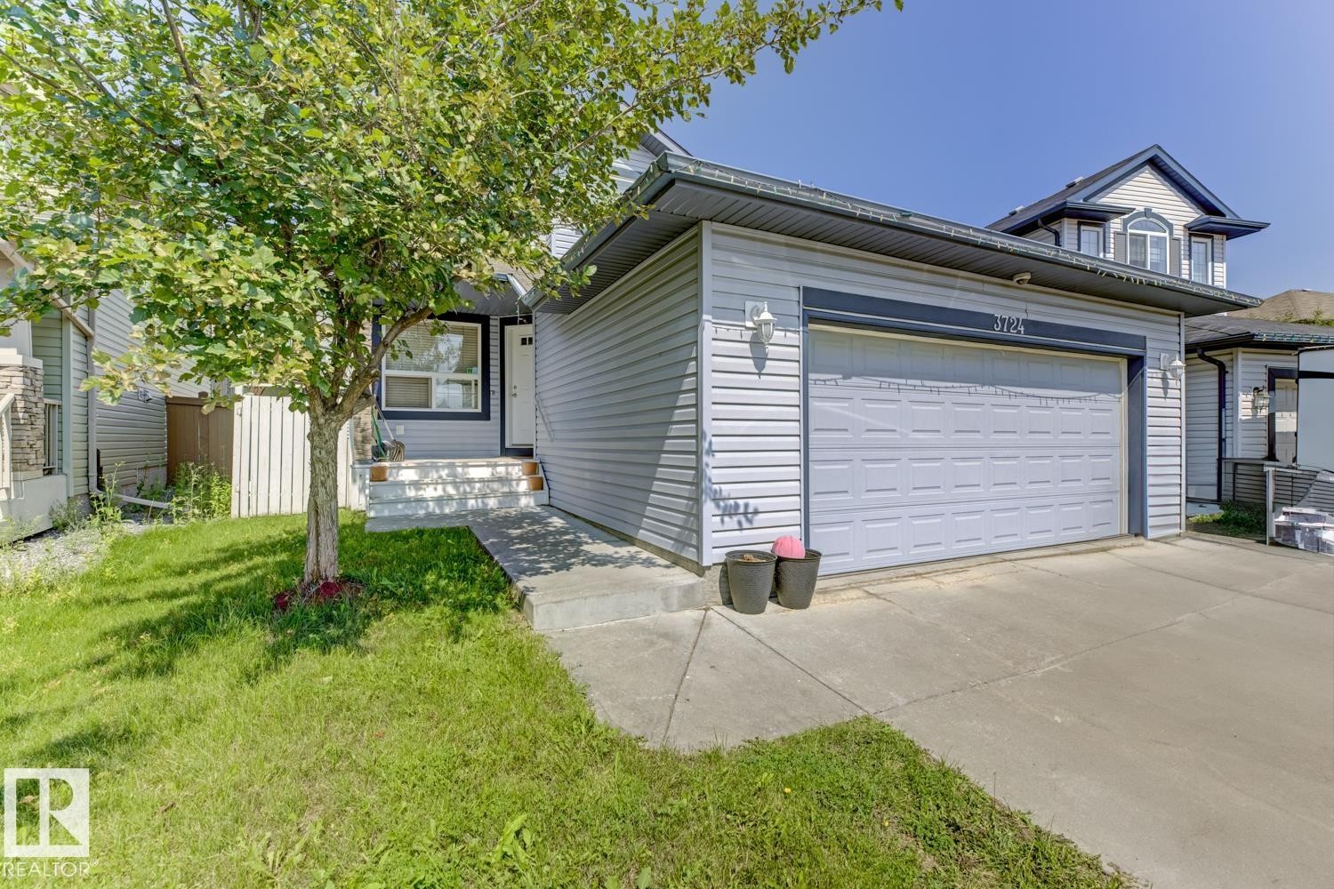 Light-toned siding exterior featuring an integrated garage with panel-style door, a concrete driveway, and a mature front yard tree - 3724 163 Avenue Nw, Edmonton, AB - Outdoor