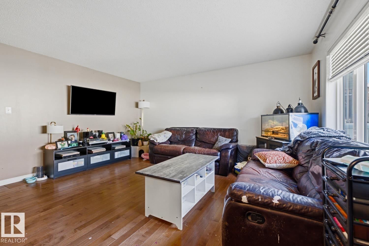Spacious living area featuring wood-finish flooring, neutral wall tones, a large window with vertical blinds, and a mounted flat-screen television - 3724 163 Avenue Nw, Edmonton, AB - Indoor Photo Showing Living Room