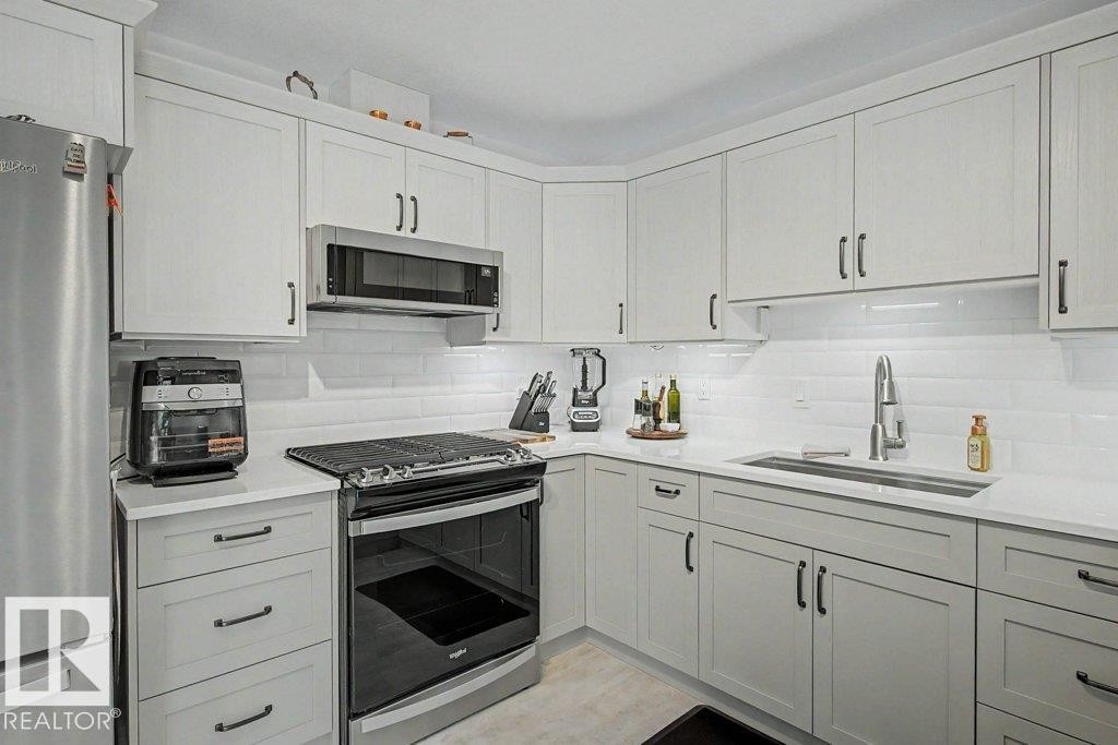 Contemporary kitchen featuring light-toned cabinetry, stainless steel appliances, a white subway tile backsplash, and a large single-basin sink with a pull-down faucet - #39 2419 133 Av Nw, Edmonton, AB - Indoor Photo Showing Kitchen With Upgraded Kitchen