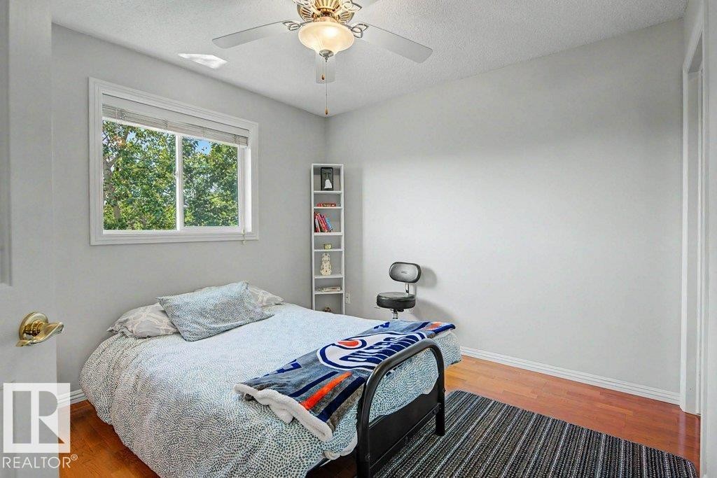 Bright room featuring a window overlooking mature trees, wood-finish flooring, a ceiling fan with integrated lighting, and a built-in shelving unit - #39 2419 133 Av Nw, Edmonton, AB - Indoor Photo Showing Bedroom