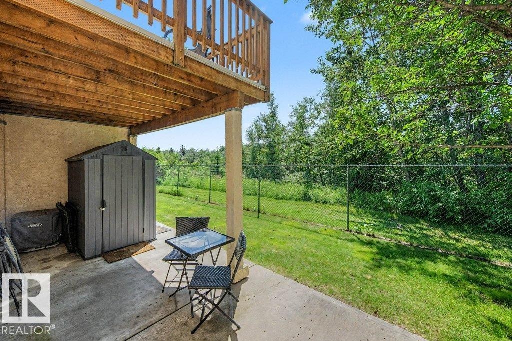 Covered concrete patio area featuring a storage shed, wood deck structure, and fenced yard with mature foliage - #39 2419 133 Av Nw, Edmonton, AB - Outdoor With Deck Patio Veranda