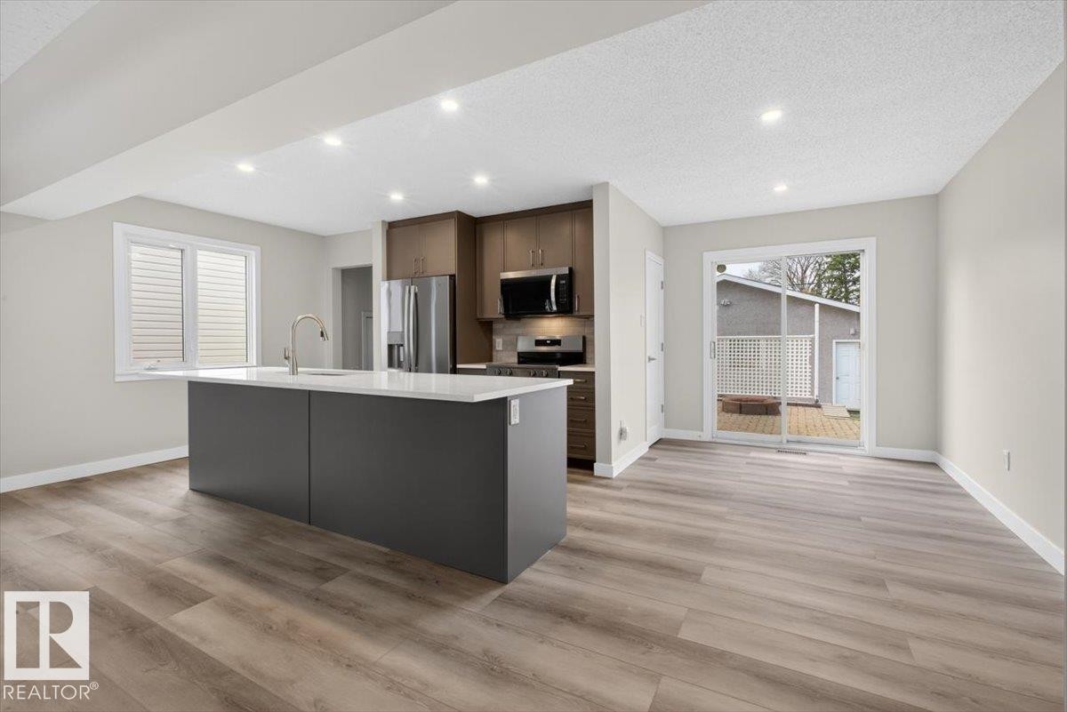 Open-concept kitchen featuring a dark gray island with a white countertop, stainless steel appliances, recessed lighting, and wood-finish flooring - 9723 65 Avenue Nw, Edmonton, AB
