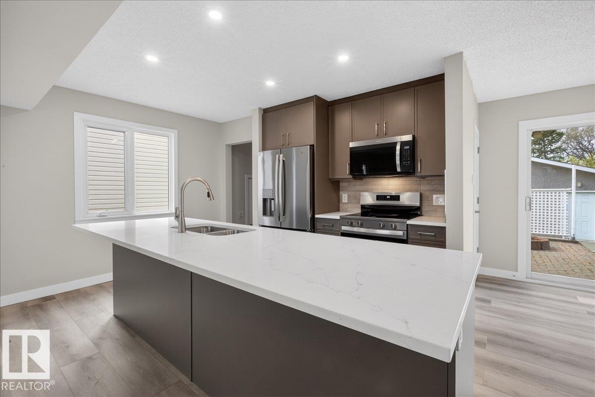 Kitchen featuring a spacious island with a white countertop and integrated sink, stainless steel appliances, contemporary cabinetry, light wood-finish flooring, and recessed ceiling lighting - 9723 65 Avenue Nw, Edmonton, AB