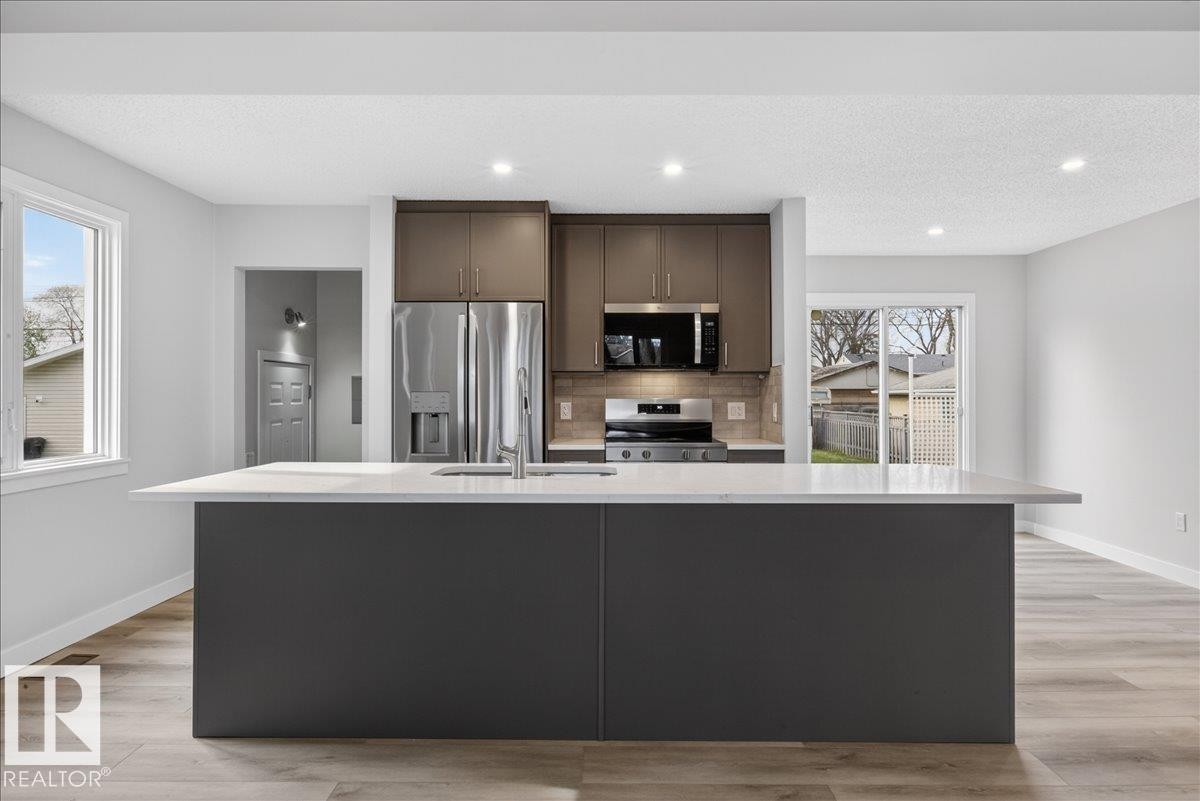 Kitchen featuring a spacious island with a white countertop, dark gray cabinetry, stainless steel appliances, and wood-finish flooring - 9723 65 Avenue Nw, Edmonton, AB