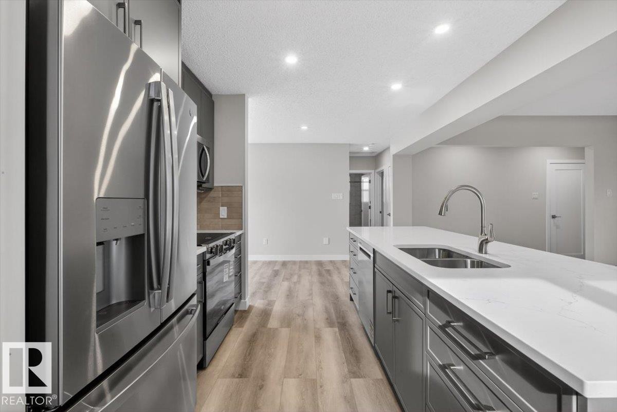 Kitchen featuring a large central island with a double basin sink and a high-arc faucet, stainless steel appliances, recessed lighting, wood-finish flooring, and shaker-style cabinetry - 9723 65 Avenue Nw, Edmonton, AB