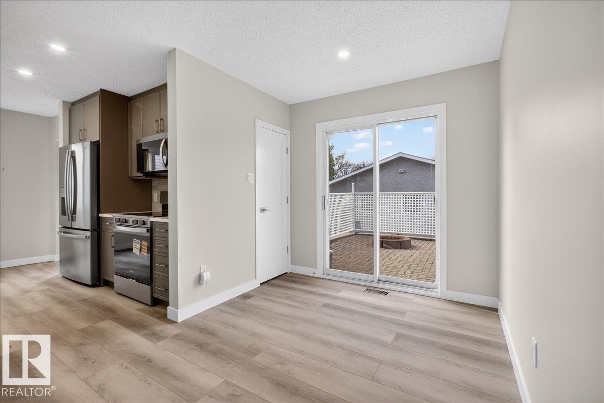 Open-concept living area with wood-finish flooring, a contemporary kitchen featuring stainless steel appliances and brown cabinetry, and a sliding glass door opening to an outdoor paver patio - 9723 65 Avenue Nw, Edmonton, AB