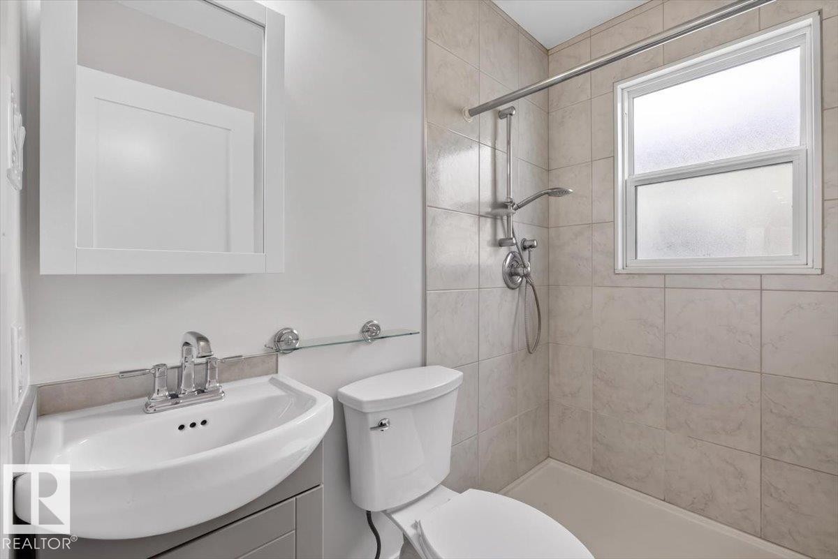 Three-piece bathroom featuring a shower with light-toned tile surround and a window, a white toilet, and a white vanity with a chrome faucet - 9723 65 Avenue Nw, Edmonton, AB