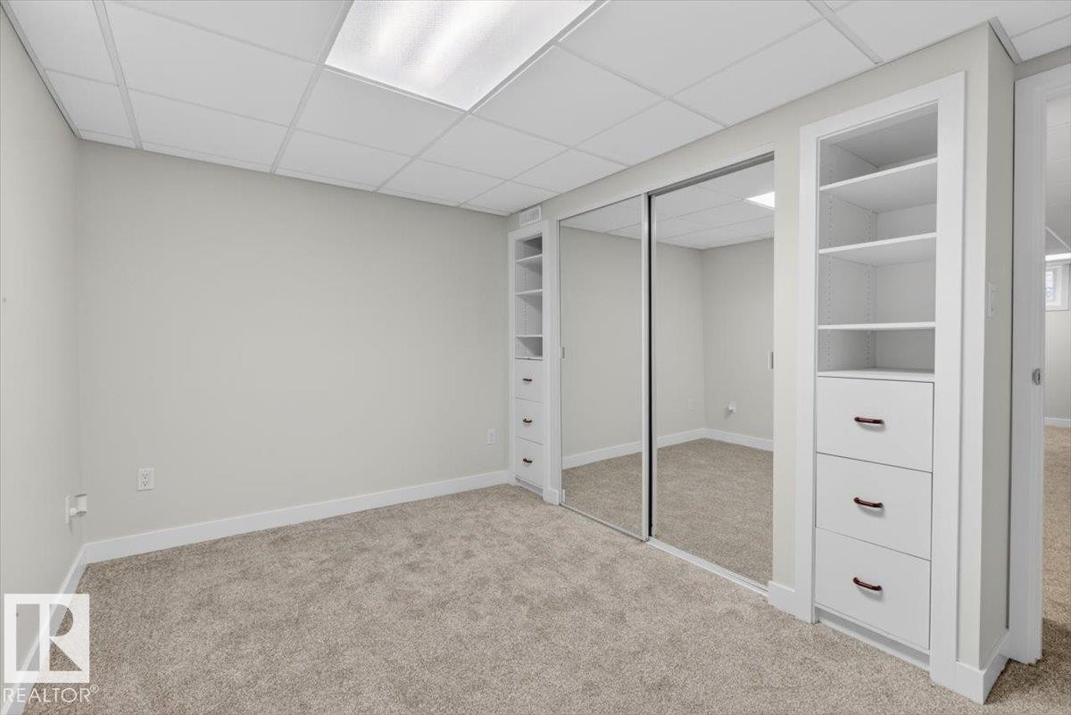 Carpeted room featuring a drop ceiling with integrated lighting, mirrored sliding closet doors, and built-in shelving units with drawers - 9723 65 Avenue Nw, Edmonton, AB