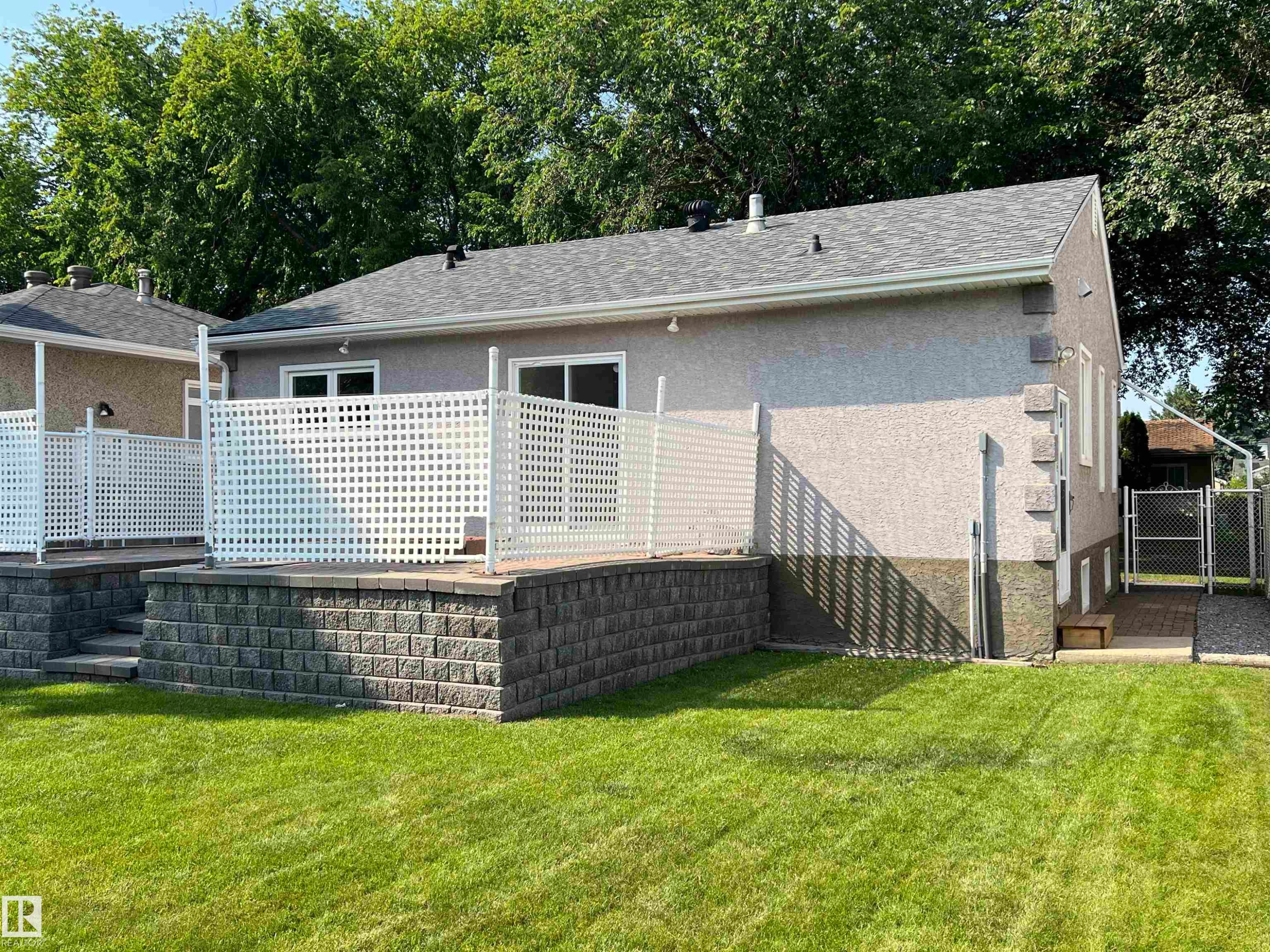 Expansive backyard featuring a manicured lawn, a raised patio with white lattice fencing, and a retaining wall constructed from stacked stone blocks - 9723 65 Avenue Nw, Edmonton, AB